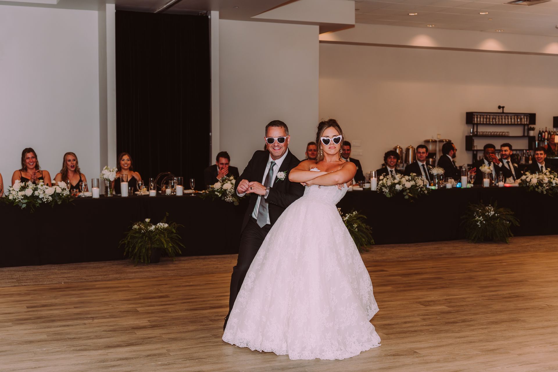 A bride and groom in sunglasses dance at their wedding reception. Guests seated at a long table in the background.