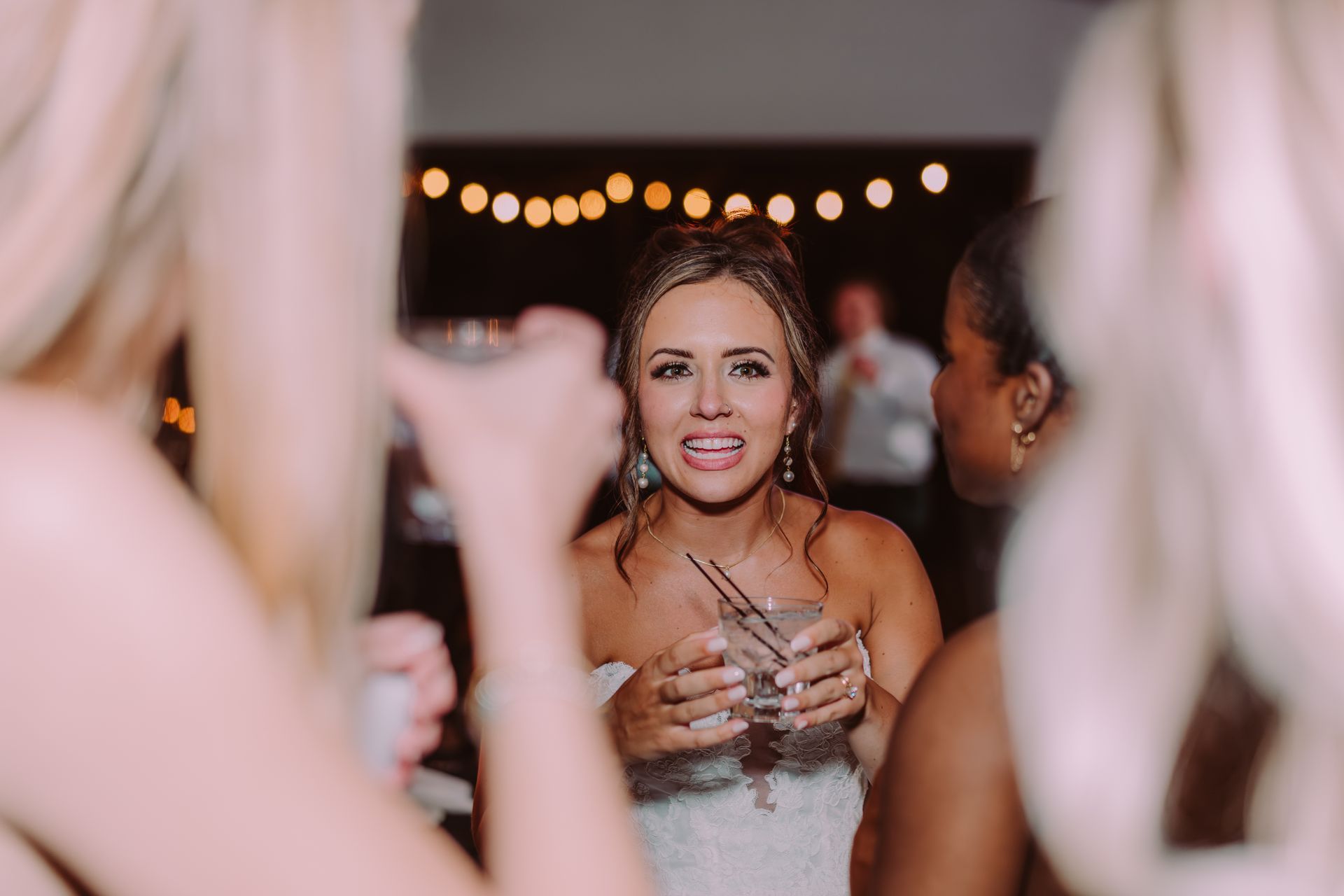 Bride with a cocktail, smiling and surrounded by bridesmaids at a wedding reception, with string lights in the background.