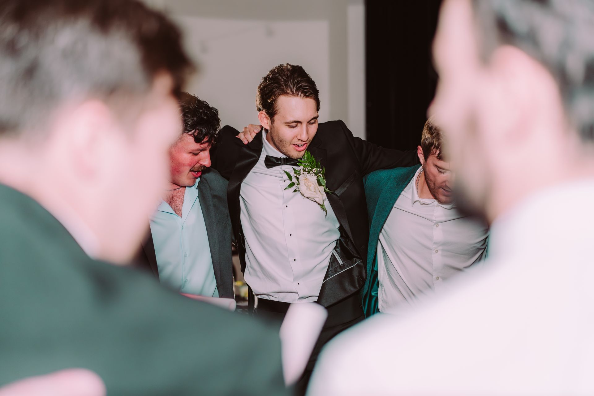 Groom surrounded by friends, adjusting his tuxedo jacket, smiling at an event. Interior setting.