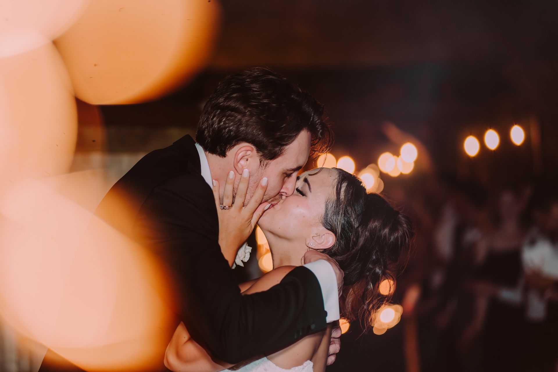 Couple kissing at a wedding reception, lit by warm fairy lights. 