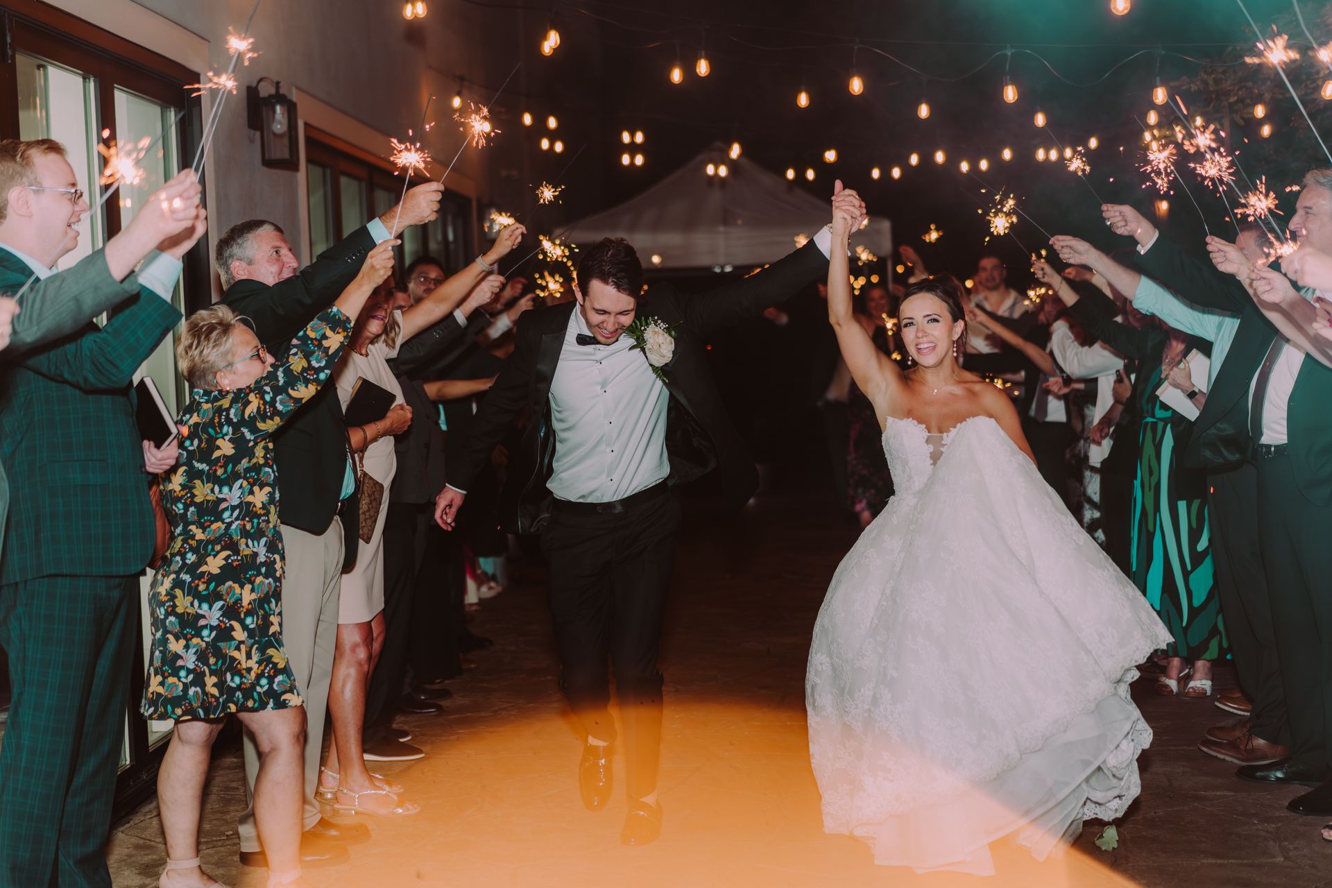 Newlyweds run through a sparkler-lit tunnel held by wedding guests, under strings of lights at an outdoor reception.