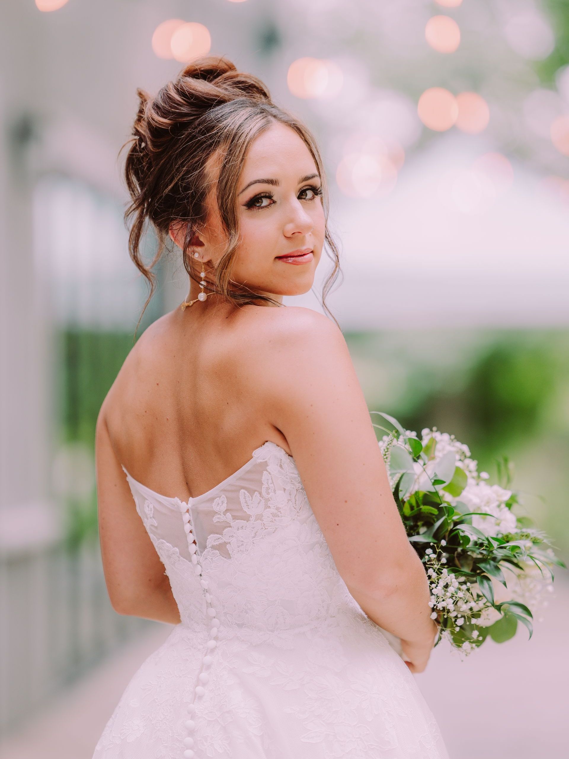 Bride in white strapless gown looks over shoulder holding bouquet; outdoor setting with blurred lights.