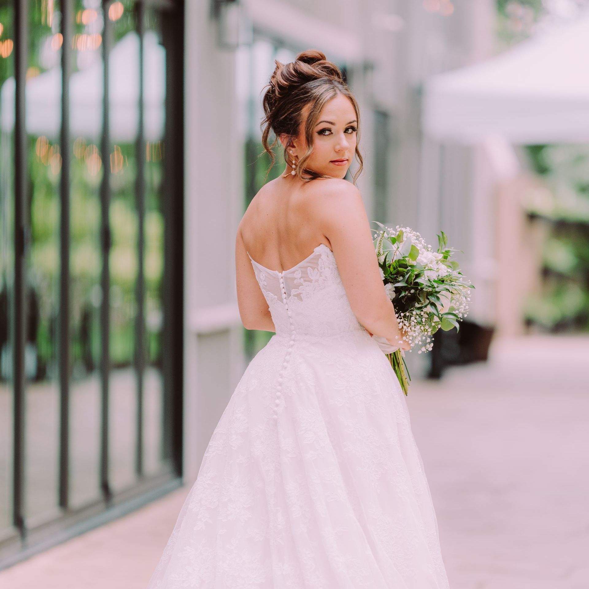 Bride in strapless white gown with updo, holding bouquet, looking over shoulder, outdoor setting.
