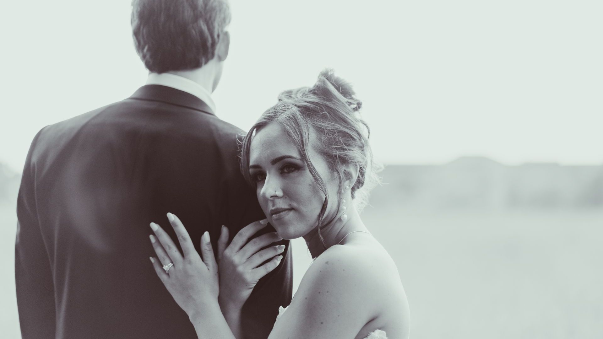 Bride resting head on groom's back, looking at the camera. Black and white, outdoors.
