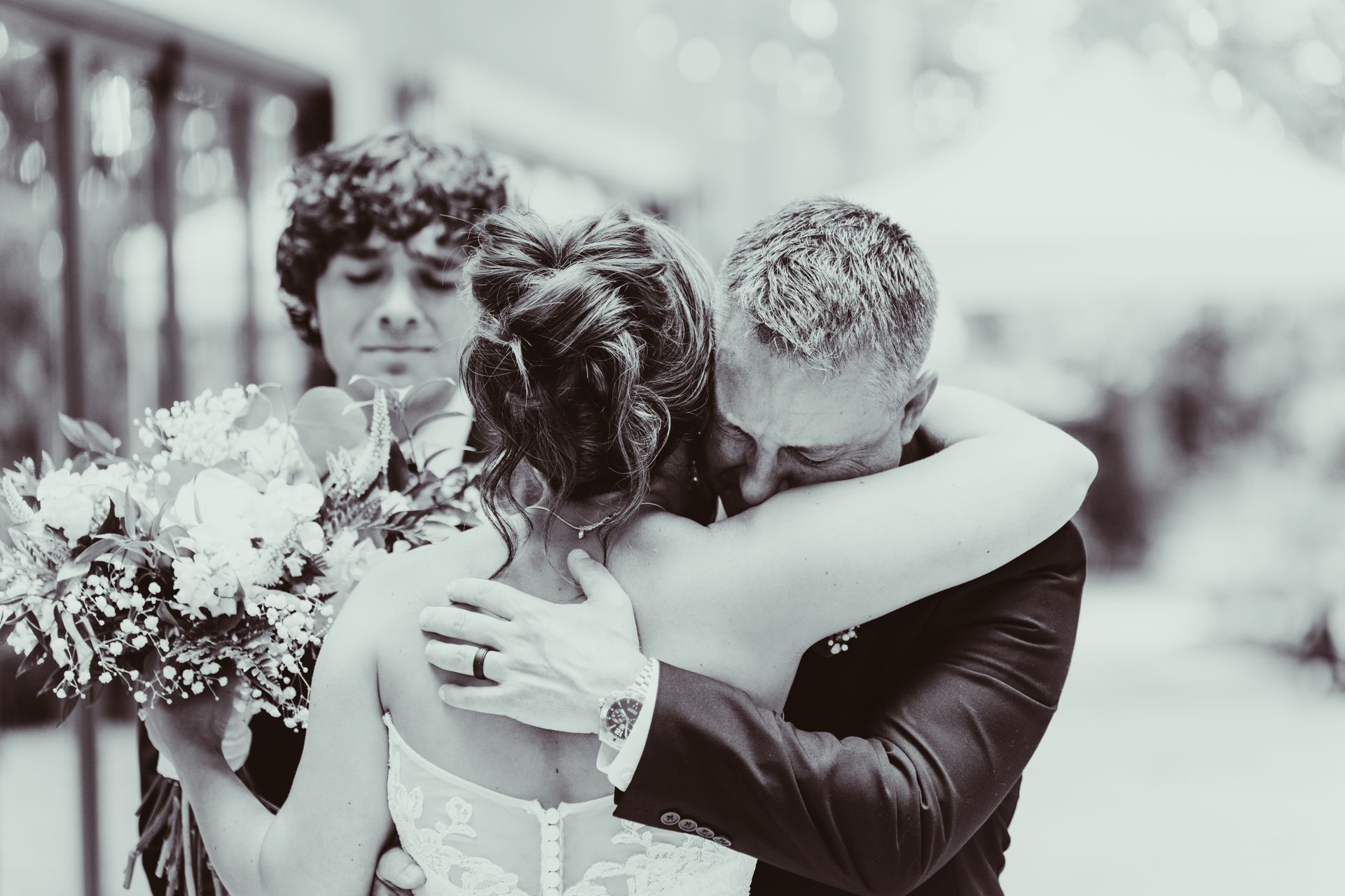 Bride hugged by two men on her wedding day. One man holds a bouquet, other embraces her.