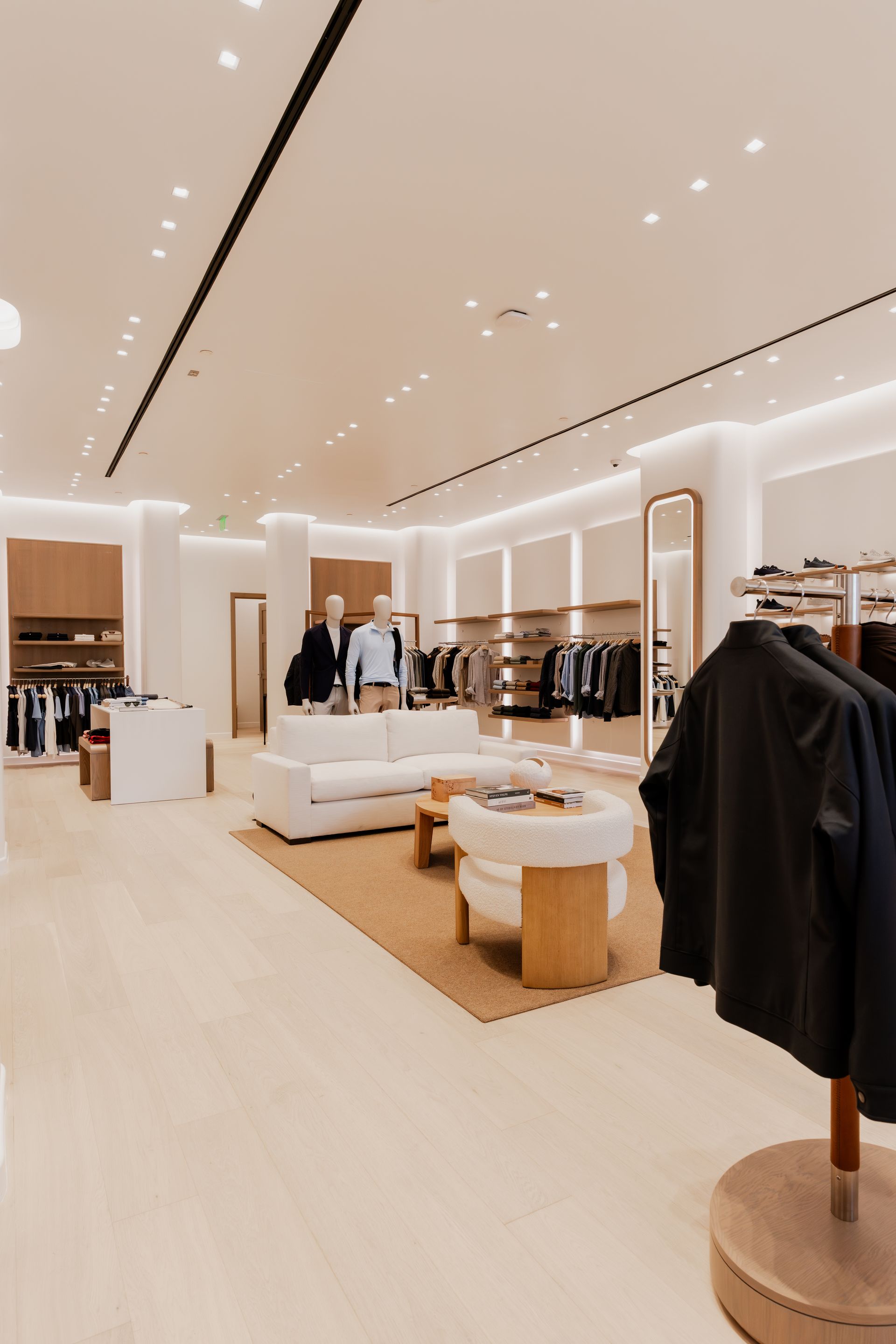 Upscale clothing store interior with clothing racks, mannequins, and seating; neutral tones.
