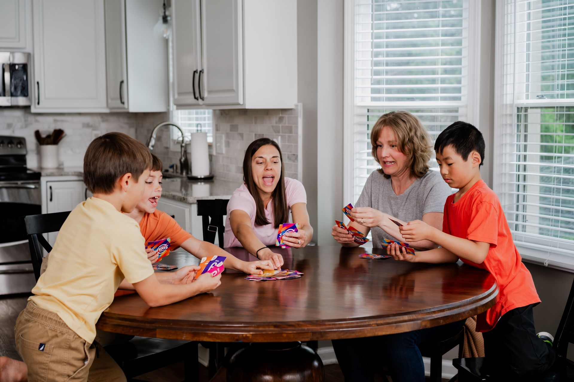 Several people sit around a dining table playing cards in a bright kitchen