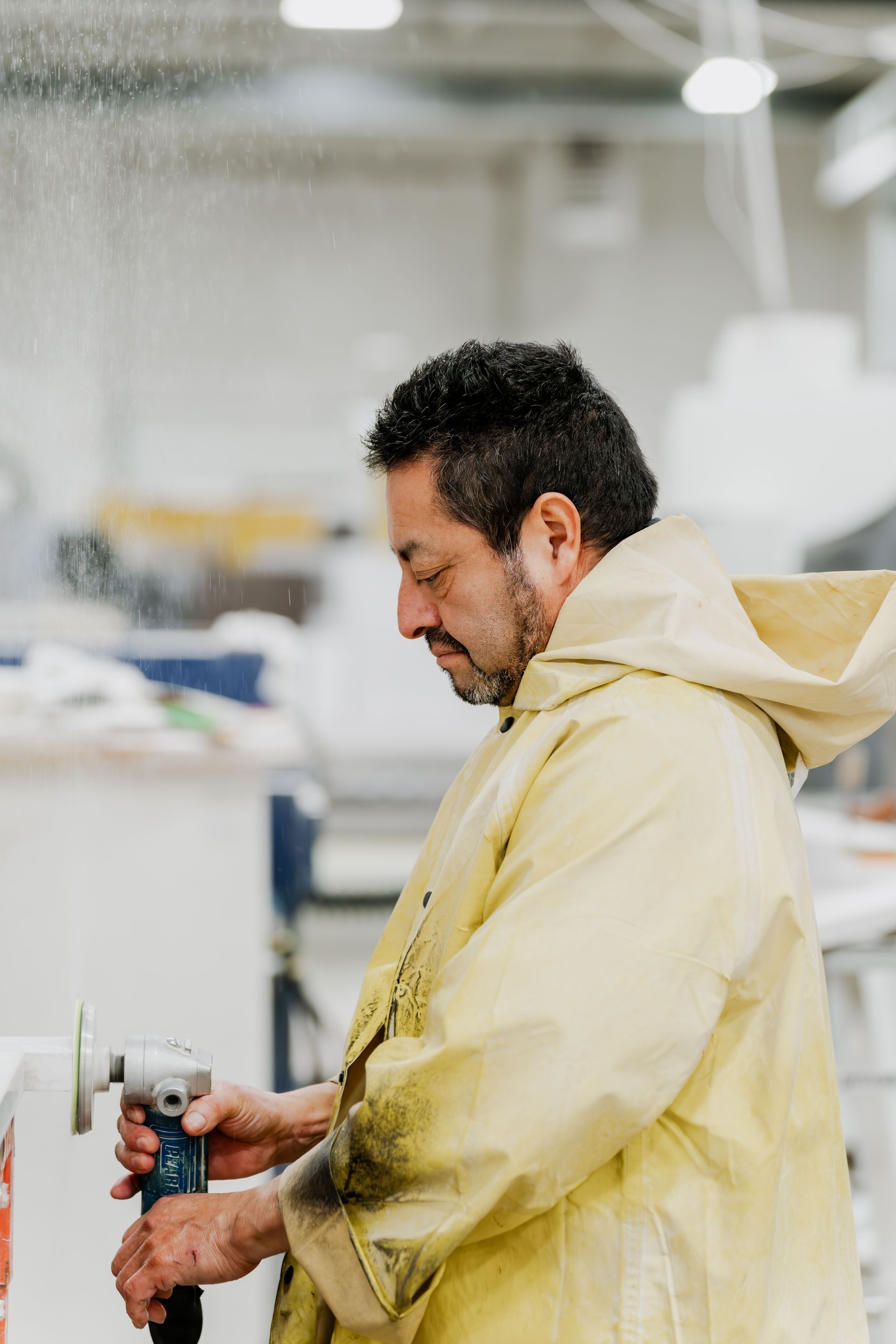 Man in yellow coat using a grinder, workshop setting, dust in air.
