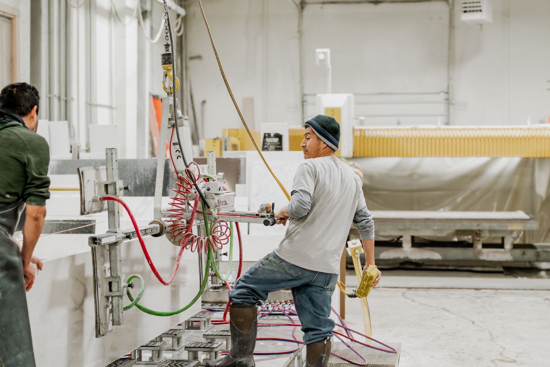 Two workers use machinery to cut stone in a workshop. One man operates the device, while the other stands nearby.