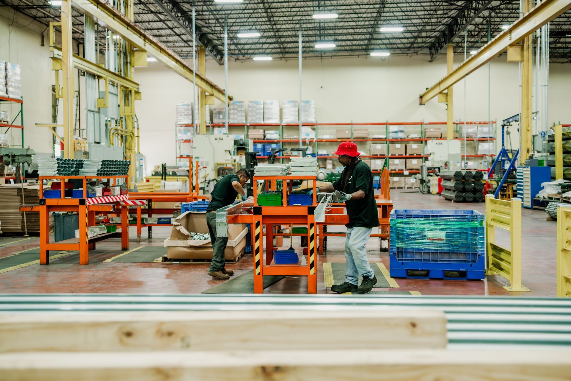Two workers in a factory assembling parts; setting includes shelving, overhead beams, and assembly tables.