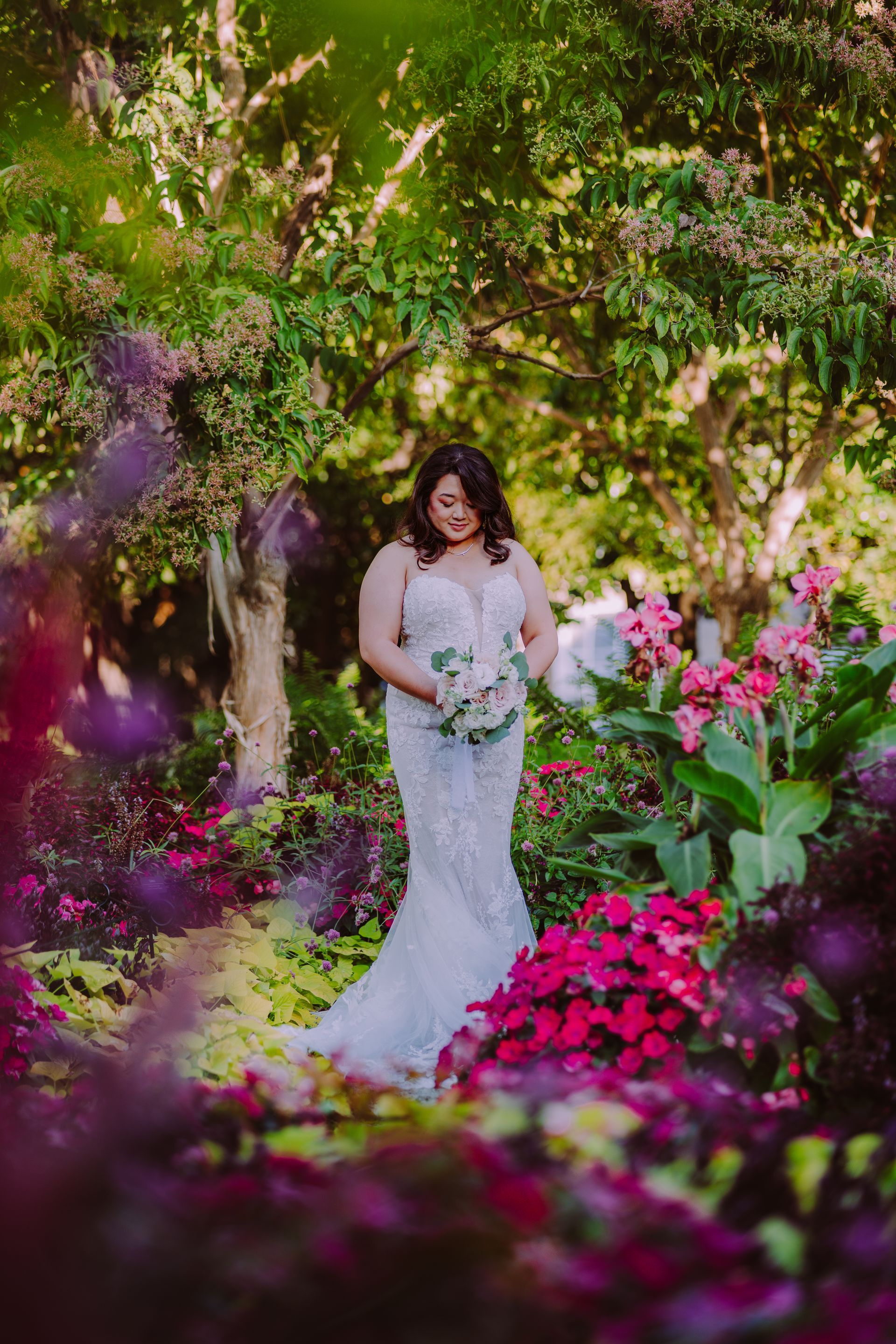 Bride in white strapless gown looks over shoulder holding bouquet; outdoor setting with blurred lights.