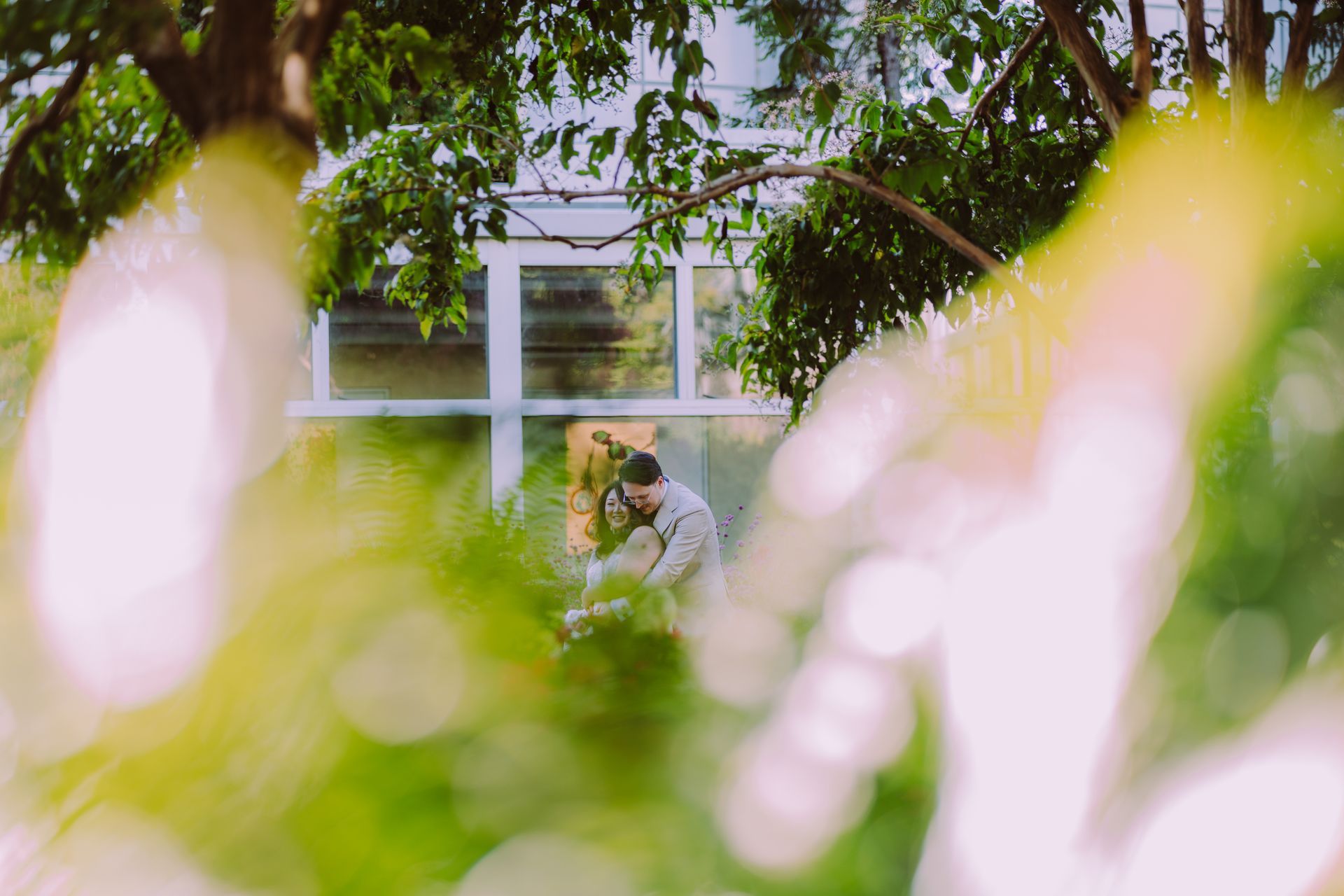 Couple embracing in front of a white-framed window, surrounded by green foliage.