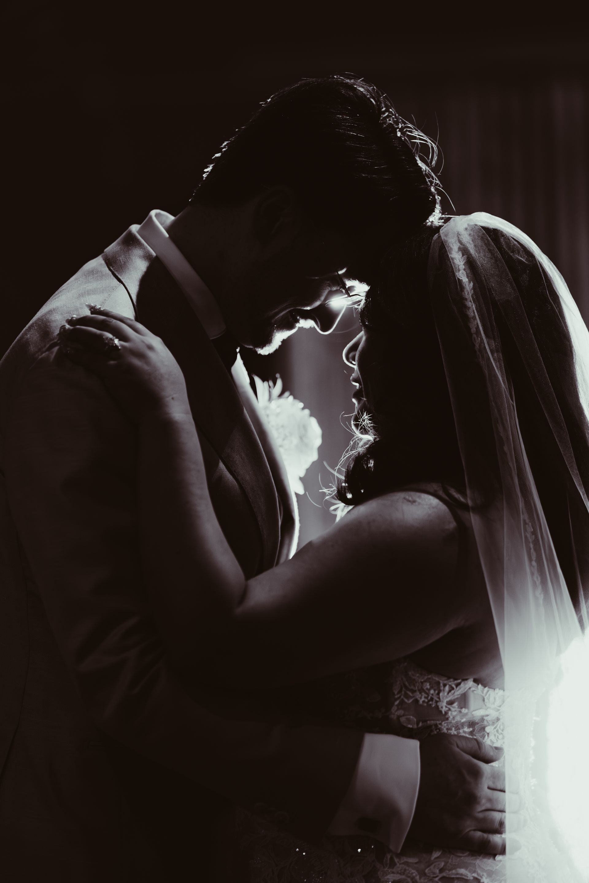 Couple in formal attire, embracing with heads close, backlit against a dark background, likely a wedding.