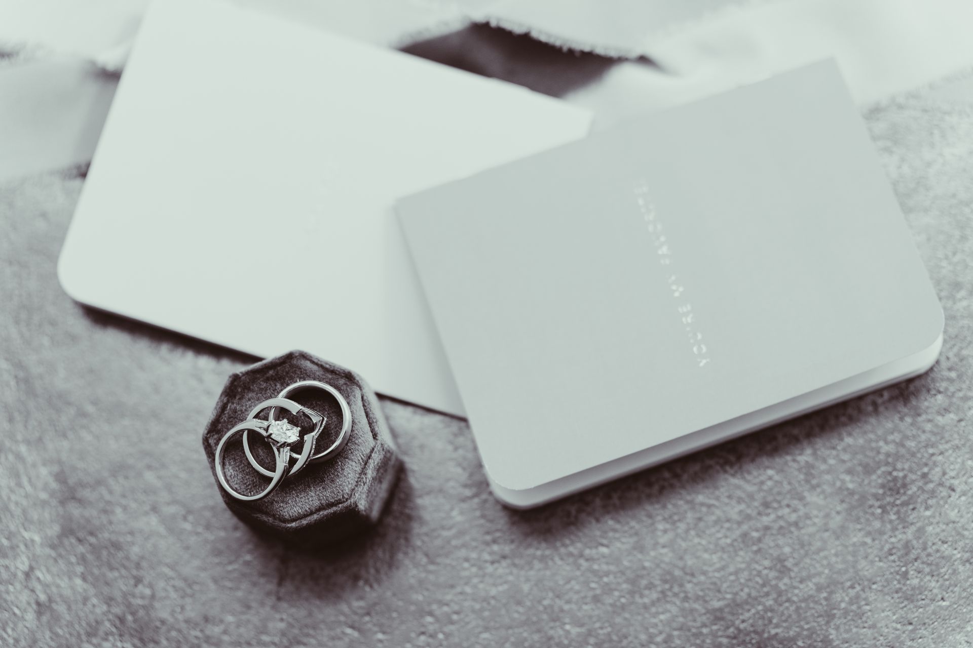 Two rings in a velvet box next to a card and an open book; all are grayscale.