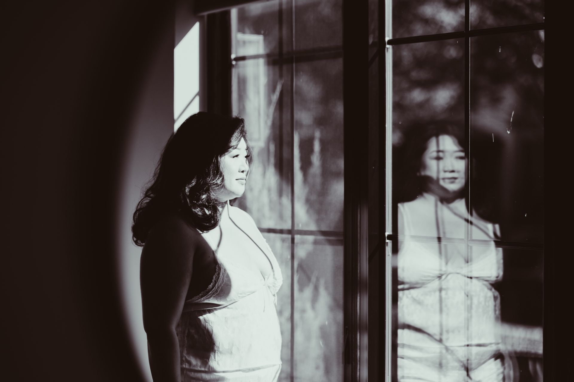 Woman standing by a window, looking out. Her reflection is visible in the glass. Black and white photo.