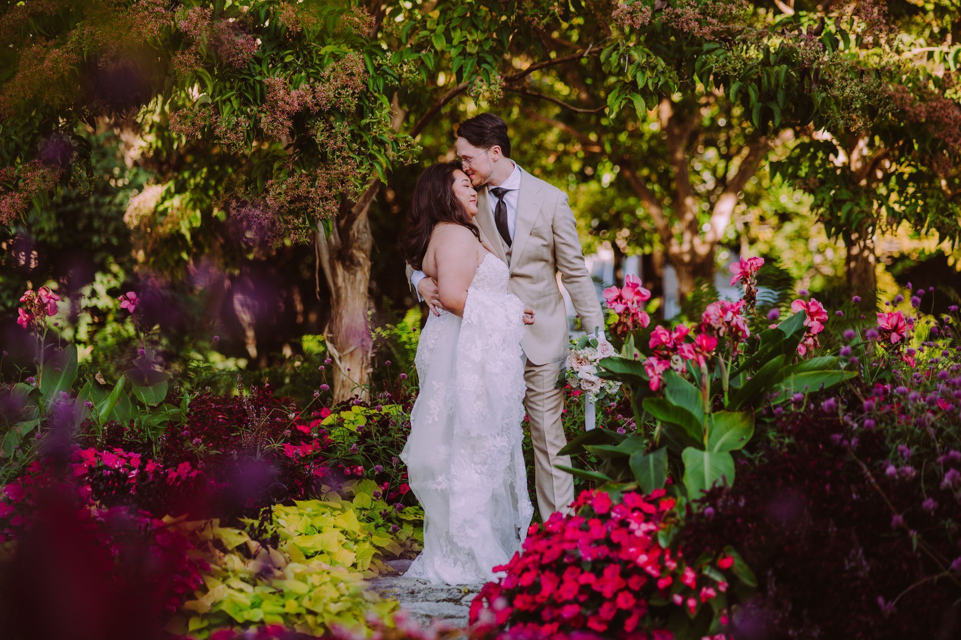 Couple embraces in a lush garden, surrounded by red and pink flowers and greenery.