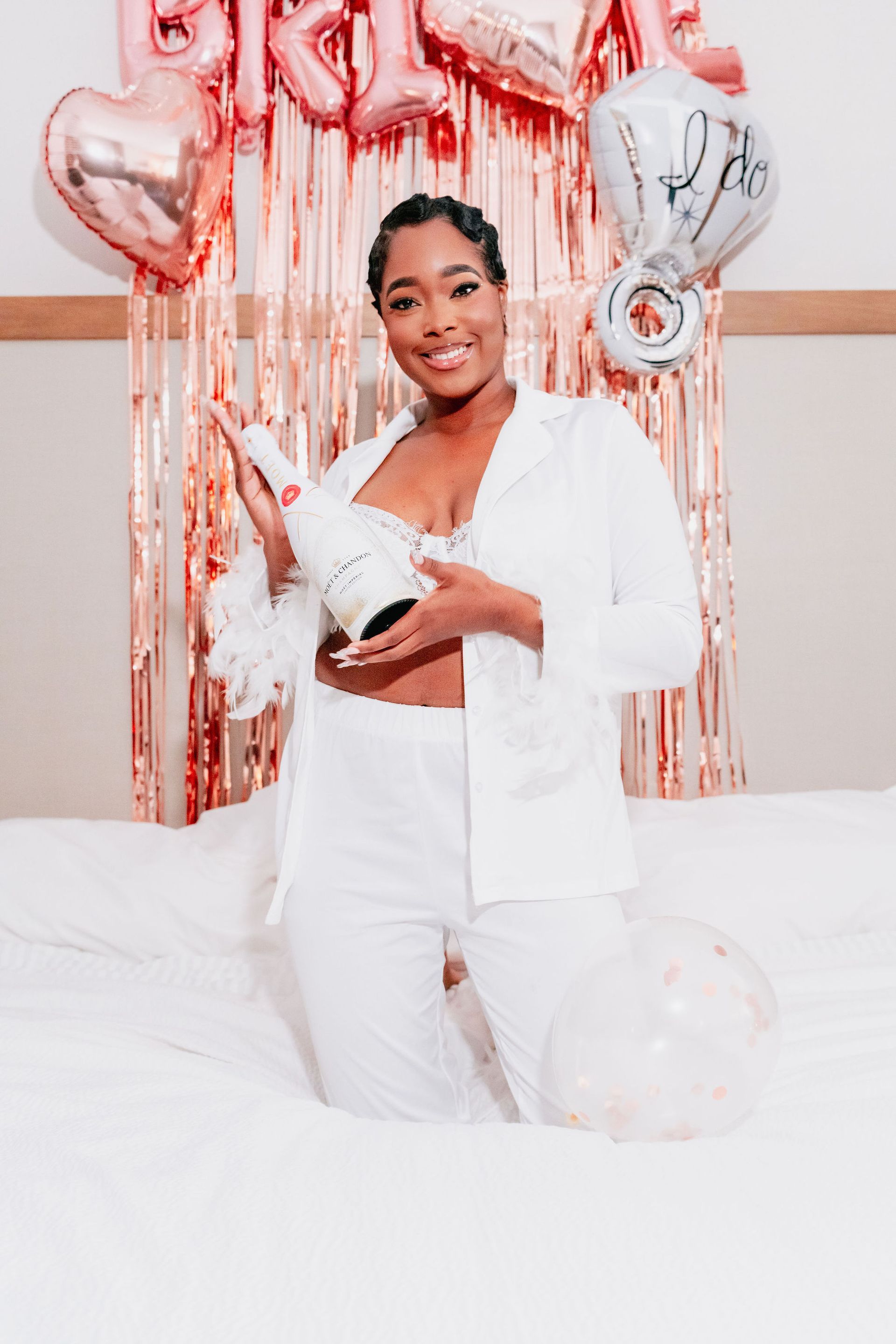 A woman in a white suit is kneeling on a bed holding a bottle of champagne.