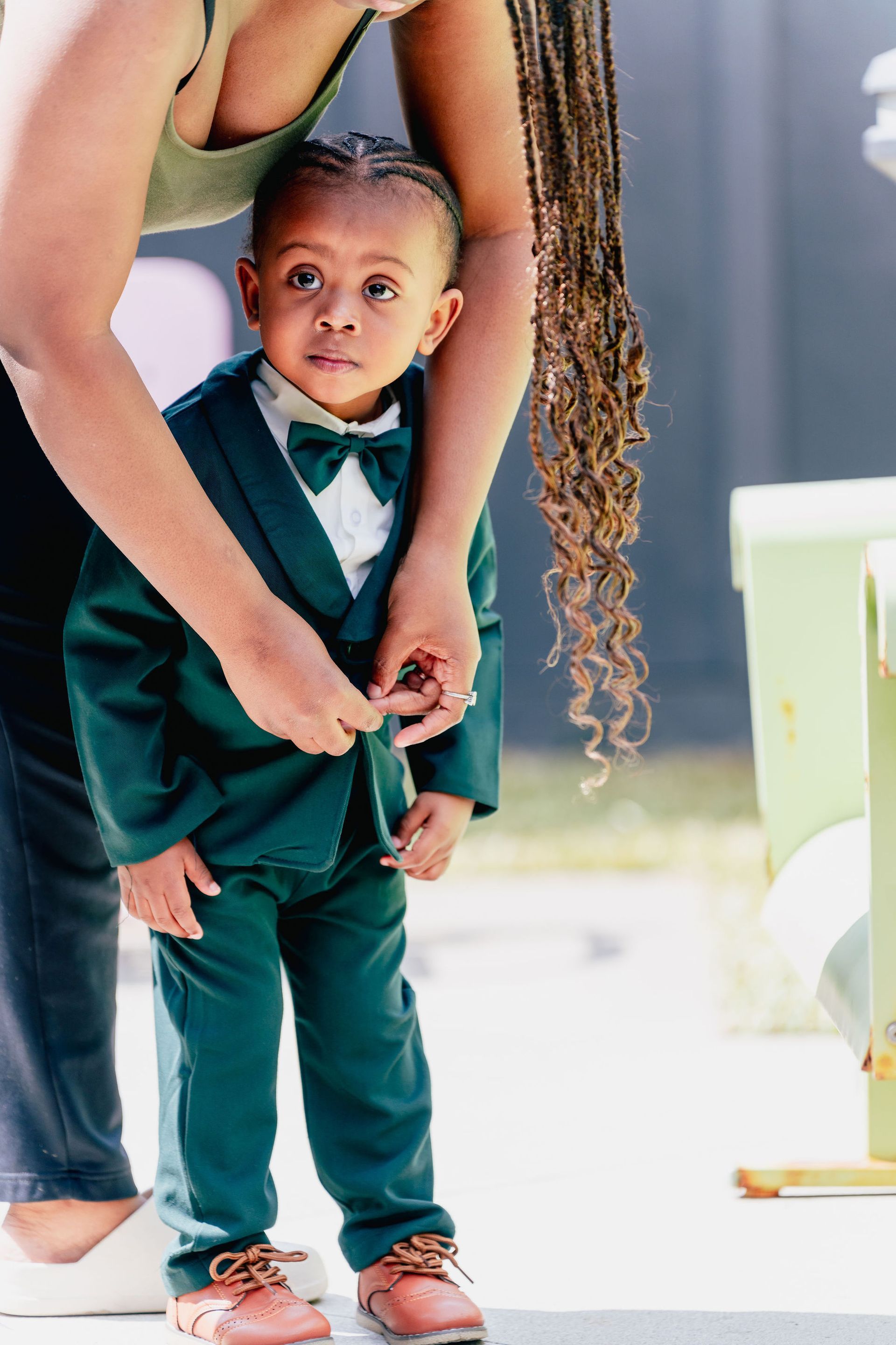 A woman is helping a little boy wear a green suit and bow tie.