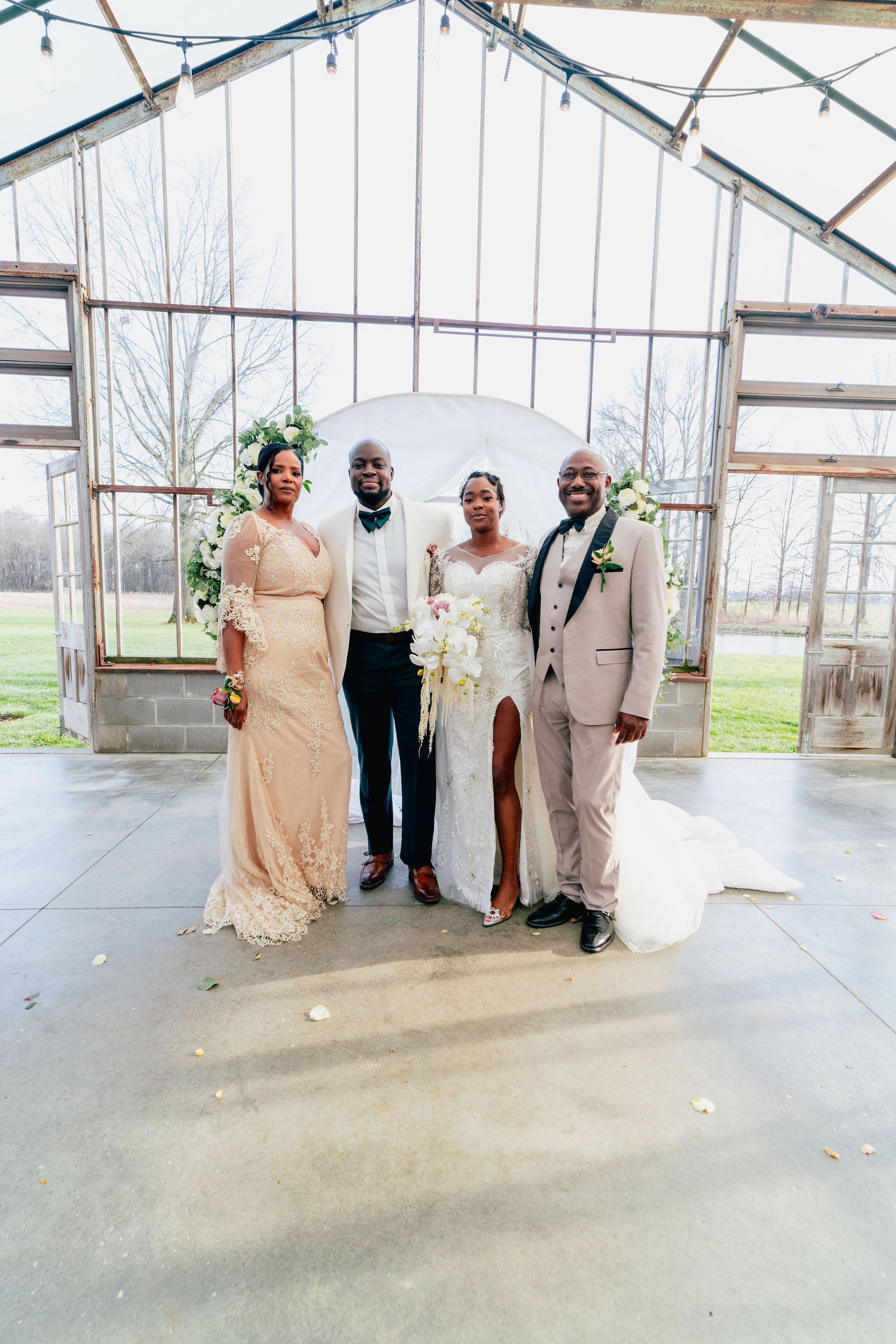 A bride and groom are posing for a picture with their parents in a greenhouse.