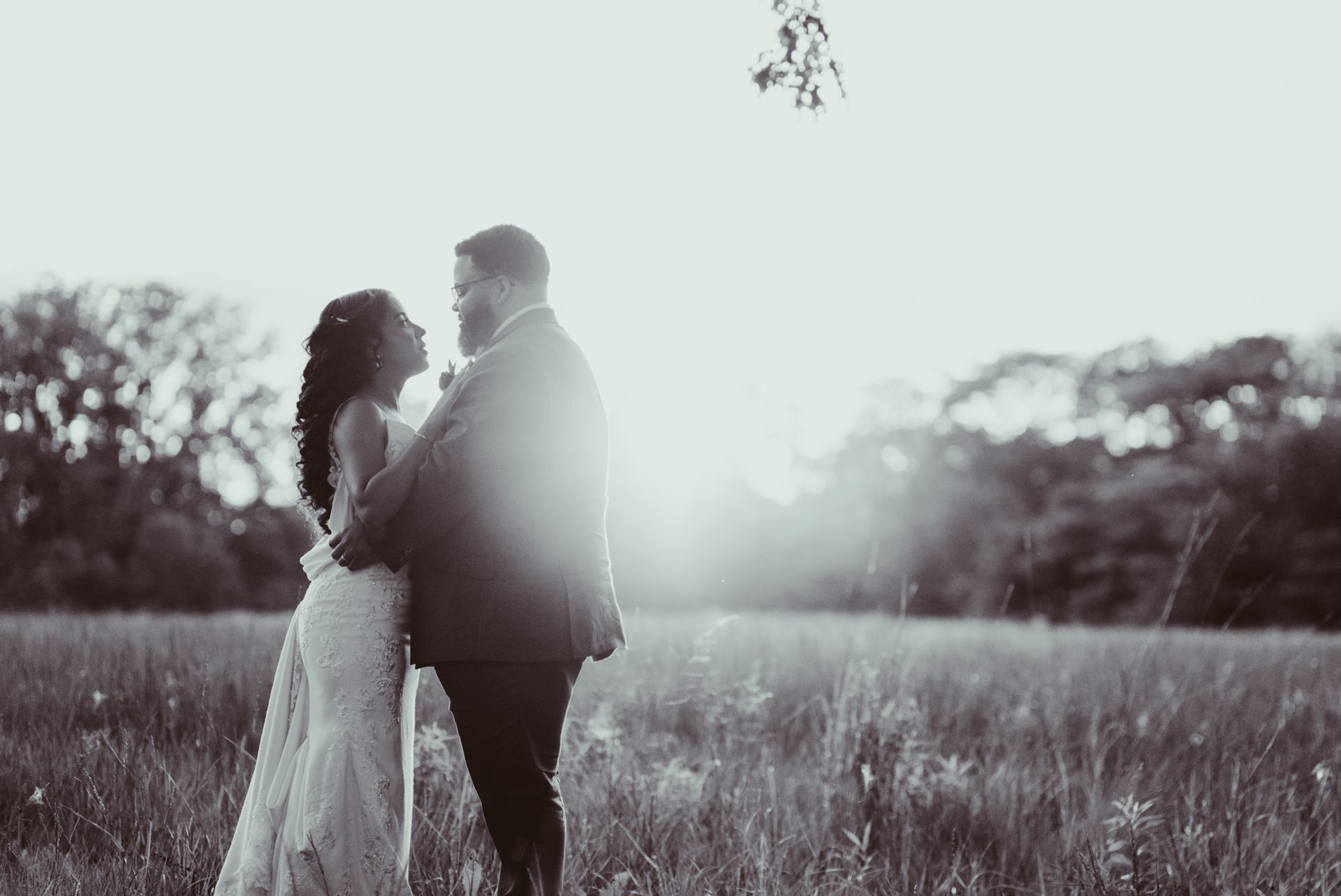 Couple embracing in field at sunset; bride in white gown, groom in suit. Black and white.