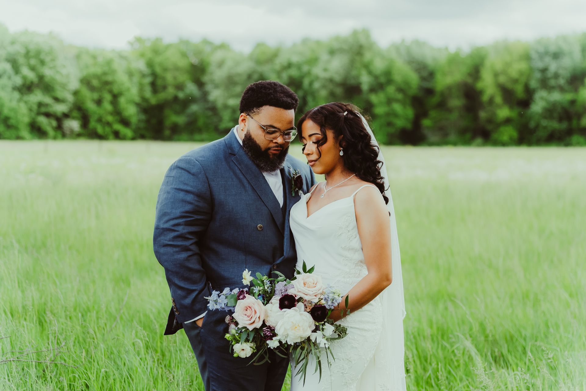 Wedding couple in field: man in blue suit, woman in white dress, holding flowers, looking down.