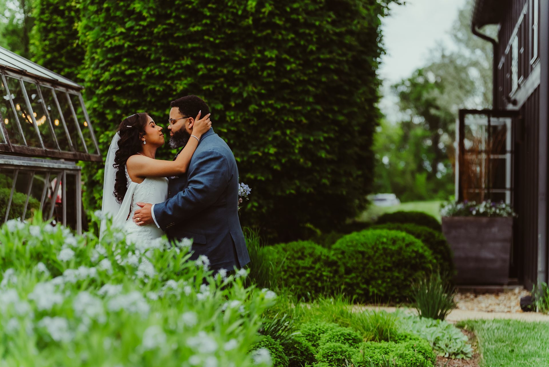 Bride and groom embrace in a garden. She wears a white dress, he a blue suit. Lush greenery surrounds them.