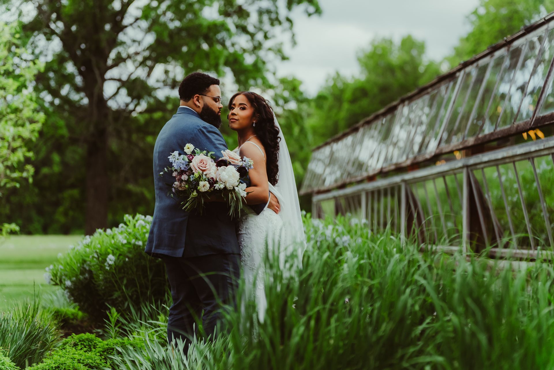 Bride and groom pose in garden near greenhouse, holding bouquet. She wears a veil. He wears a blue suit.