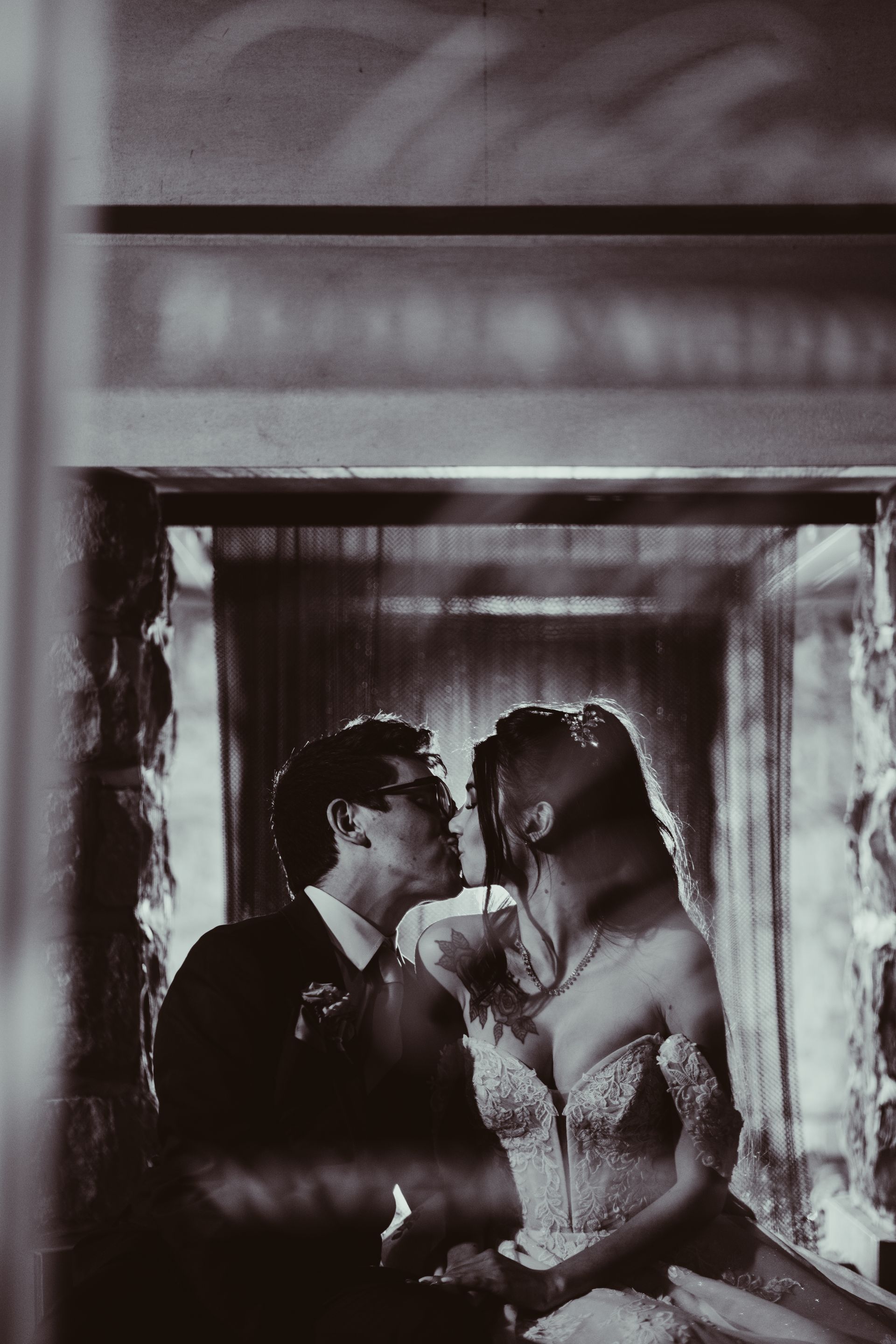 Bride and groom in embrace, silhouetted in a window, backlit. Wedding dress, suit, kissing. Black and white.