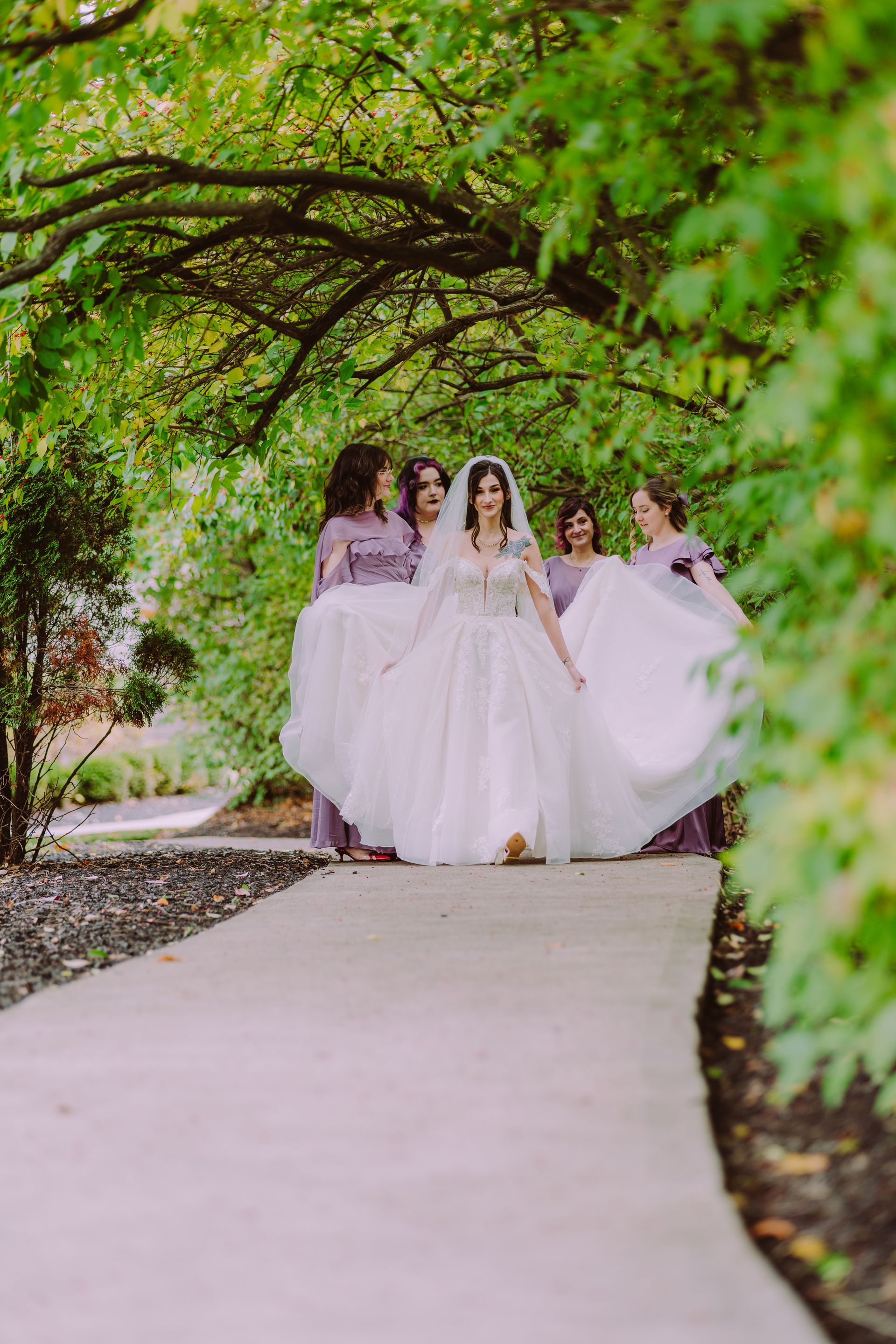 Bride in wedding dress, walking with bridesmaids along a path under a tree canopy at watersedge event center.