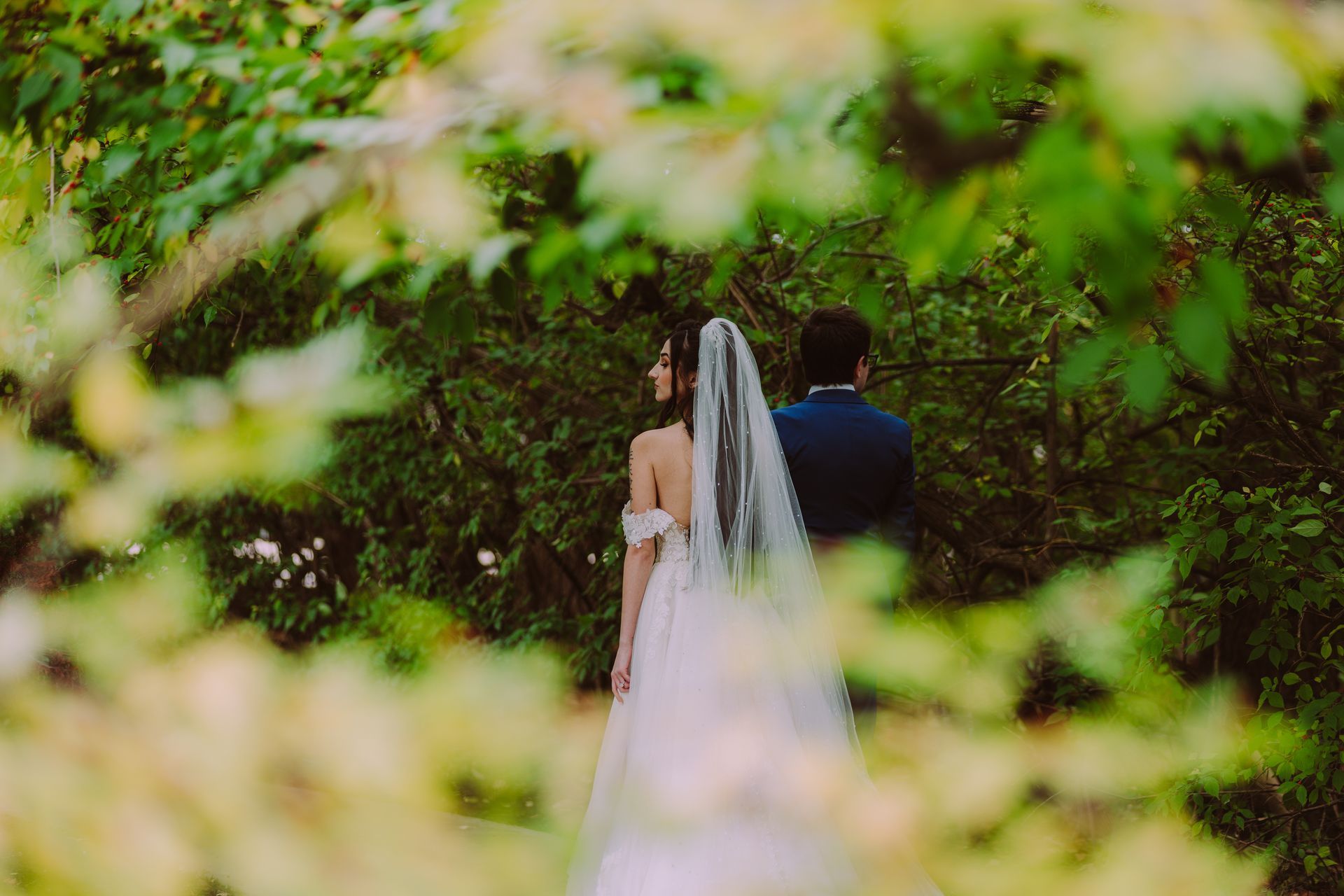 Bride and groom in formal wear stand in a lush green forest at watersedge event center.