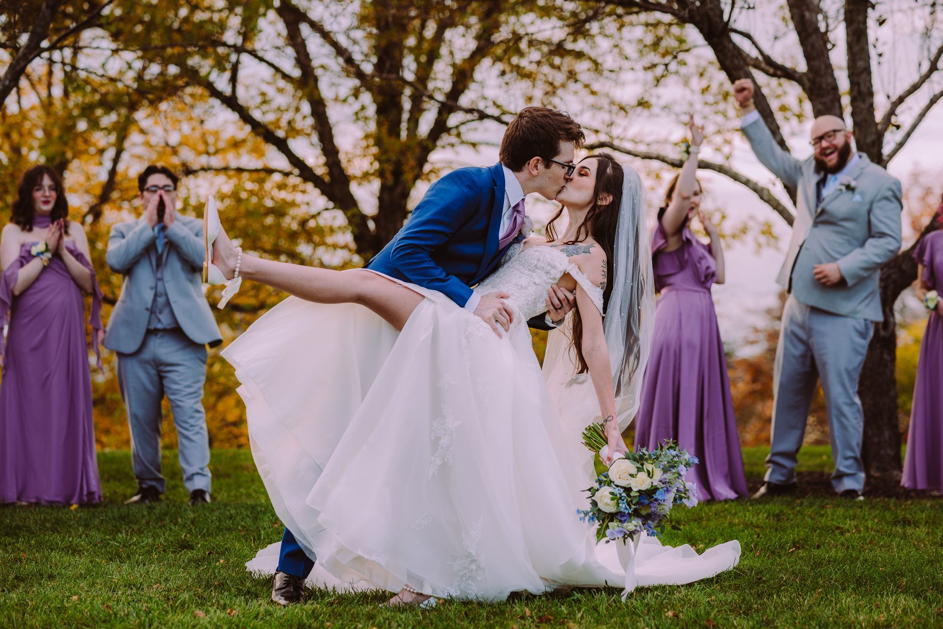 Bride and groom kissing, groom dipping bride. Wedding party cheers in a grassy outdoor setting at watersedge event center.