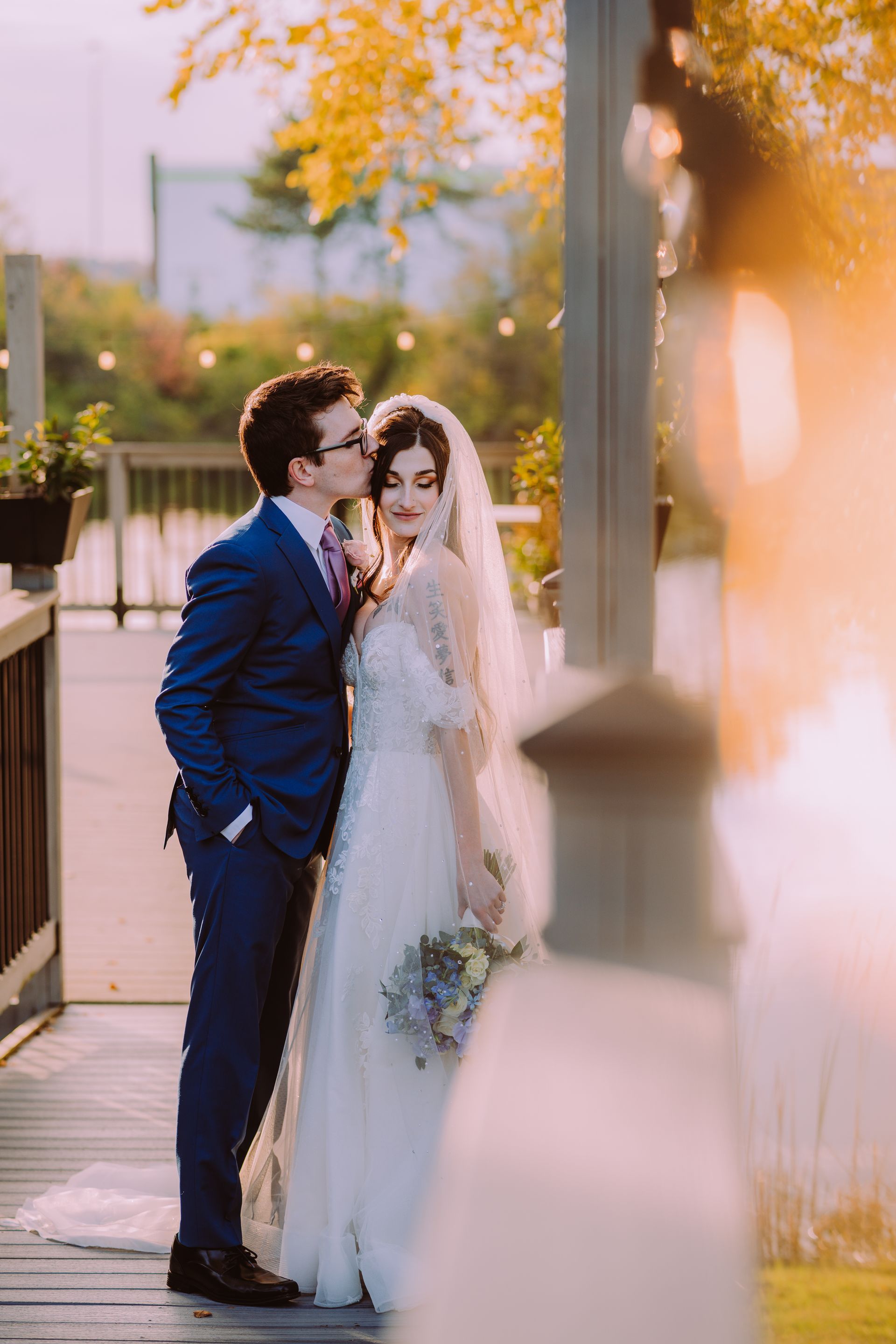 Groom kisses bride's forehead on a wooden dock at sunset at watersedge event center.