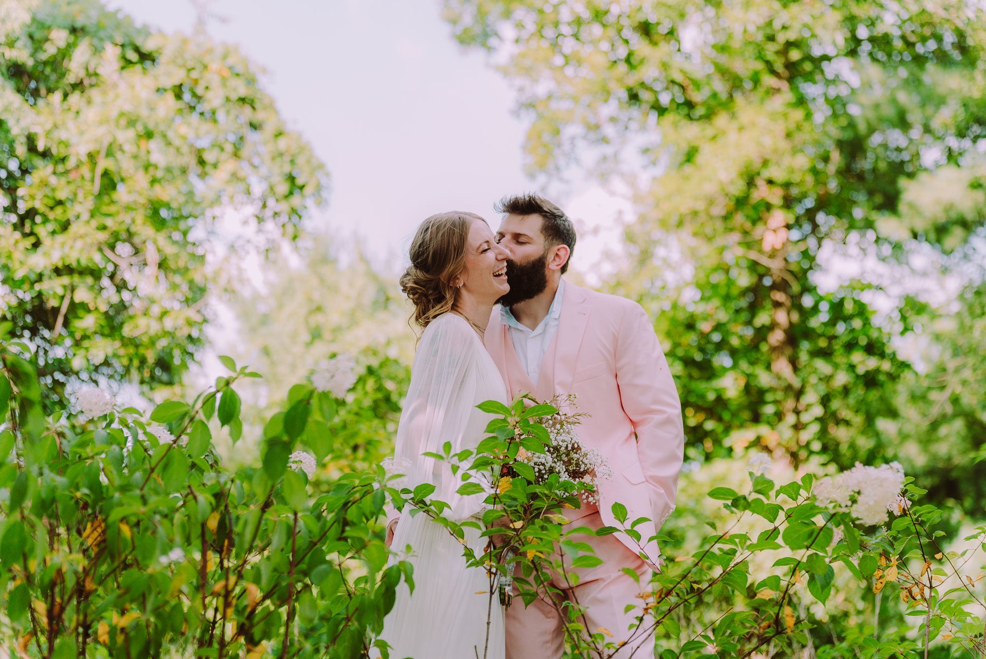 Couple in wedding attire embrace outdoors among greenery; man in pink suit kisses woman's cheek.
