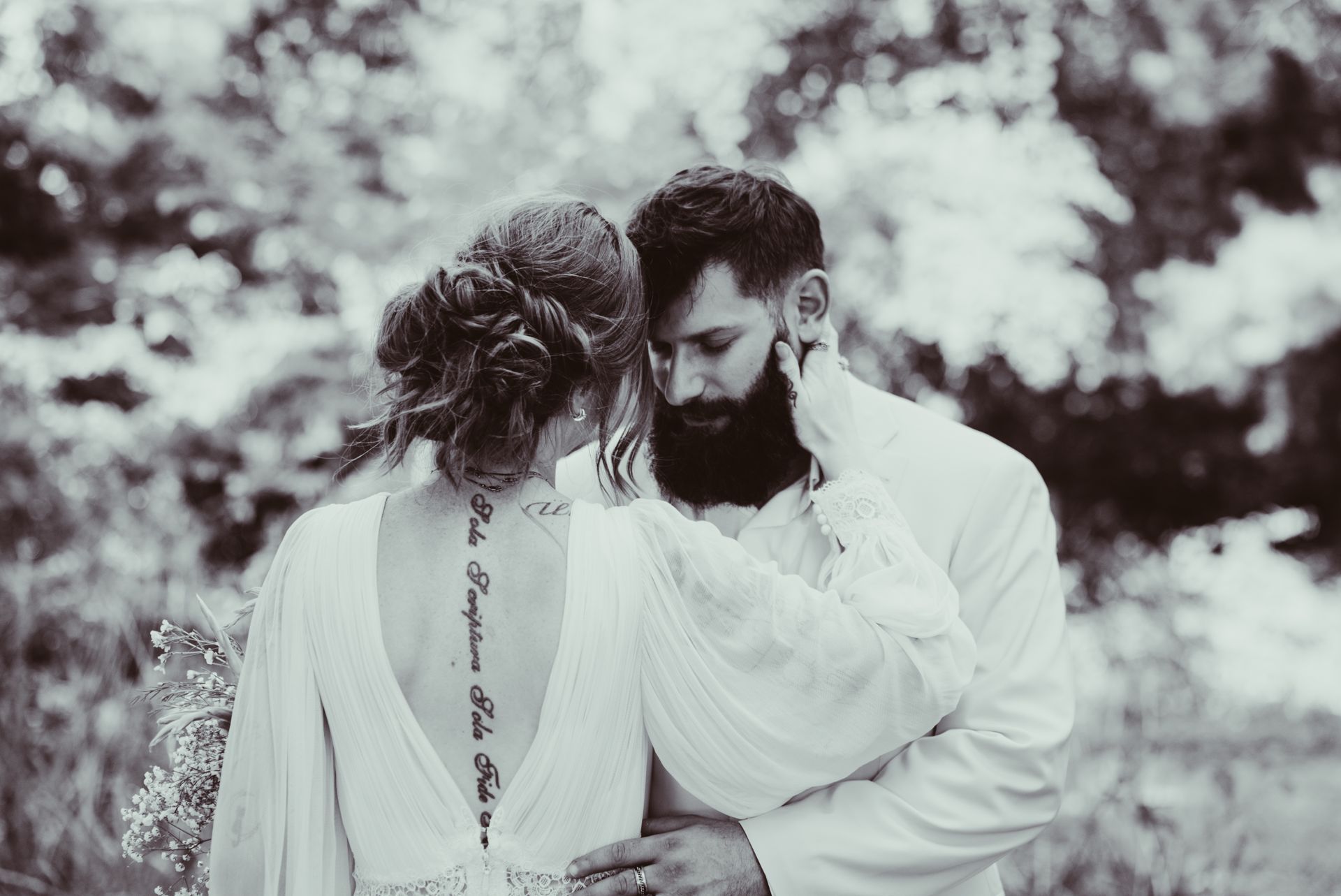 Bride and groom embrace outdoors; she wears a dress with back tattoo, he has a beard.