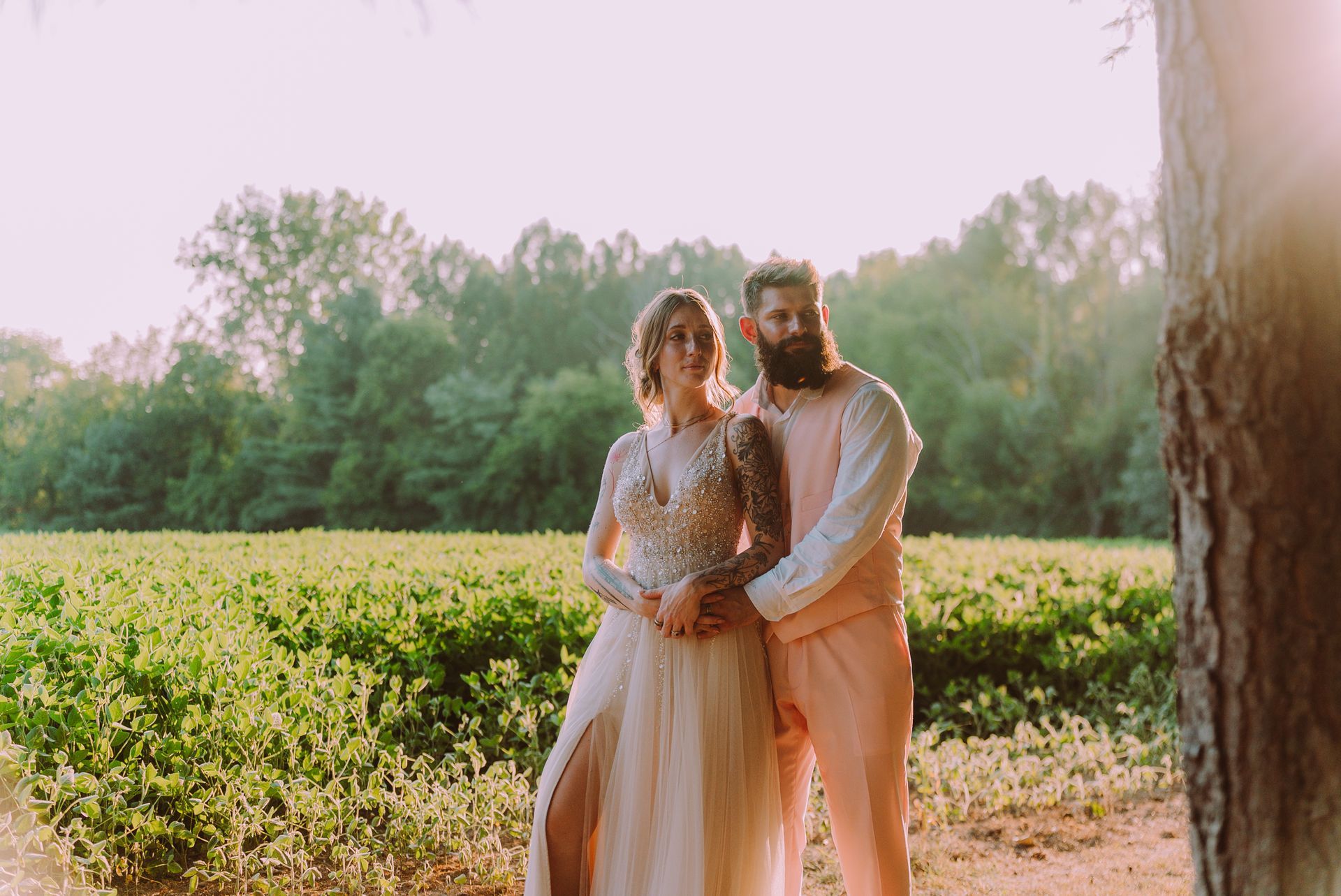 Couple in pastel outfits embrace in a sunlit field; trees in background.