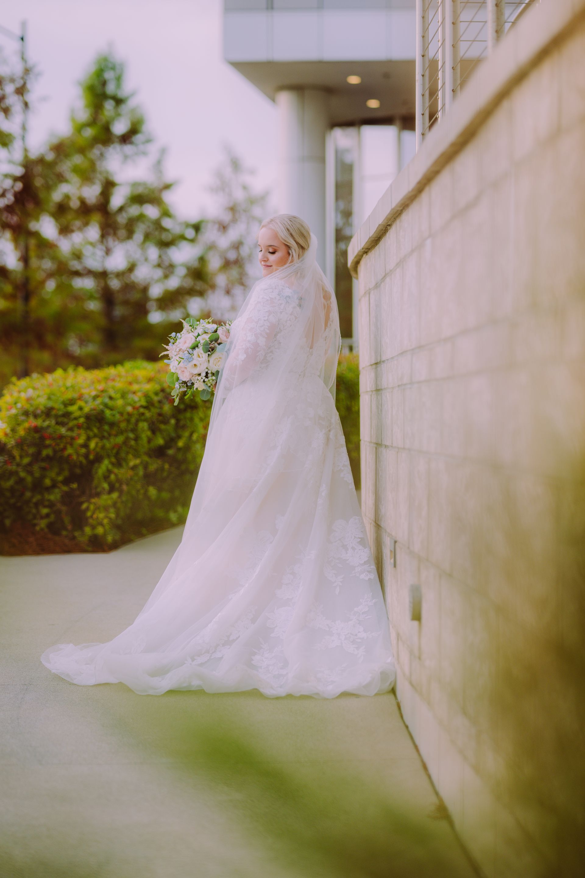 Bride in a white wedding dress and veil holding flowers, leaning against a stone wall.