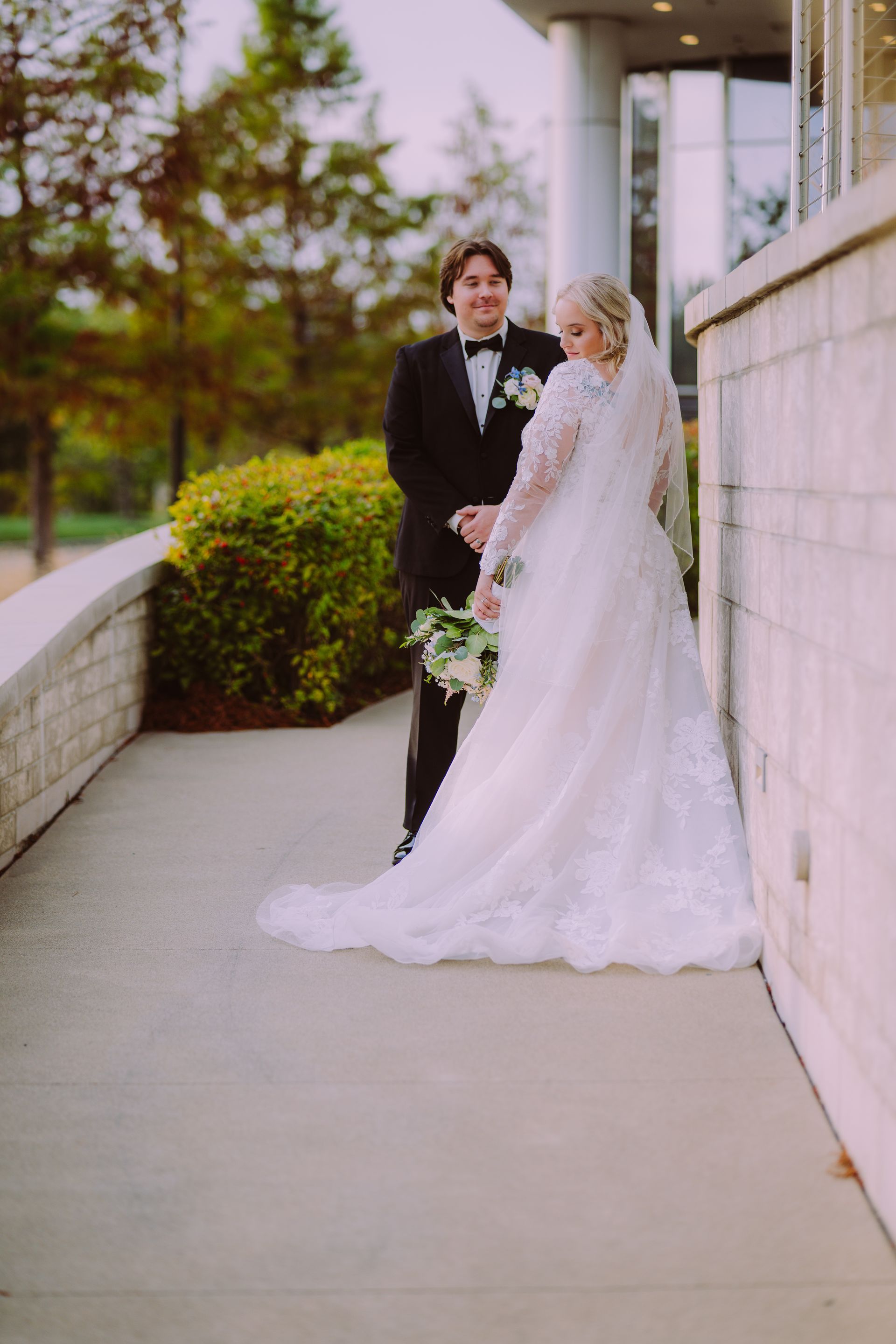 Bride and groom pose outside a building. The bride wears a white gown and veil, the groom a black tuxedo.