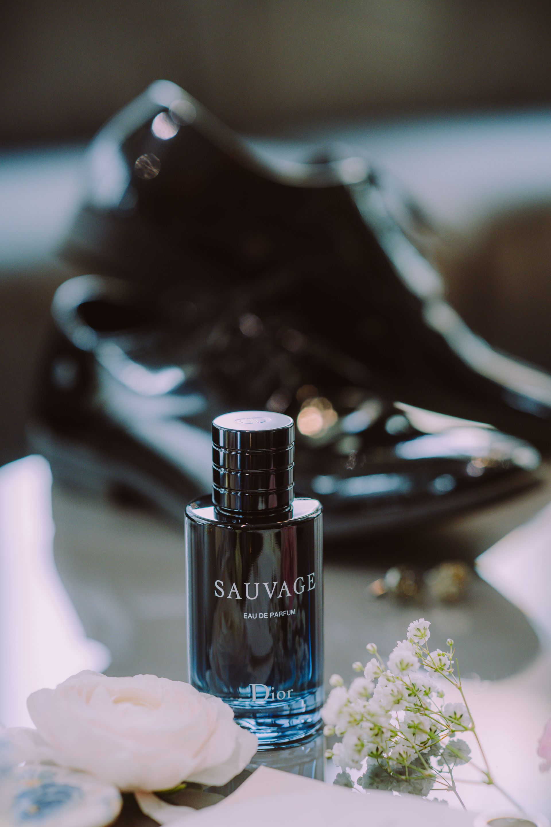 Salvage perfume bottle next to black dress shoes and white flowers on a reflective surface.