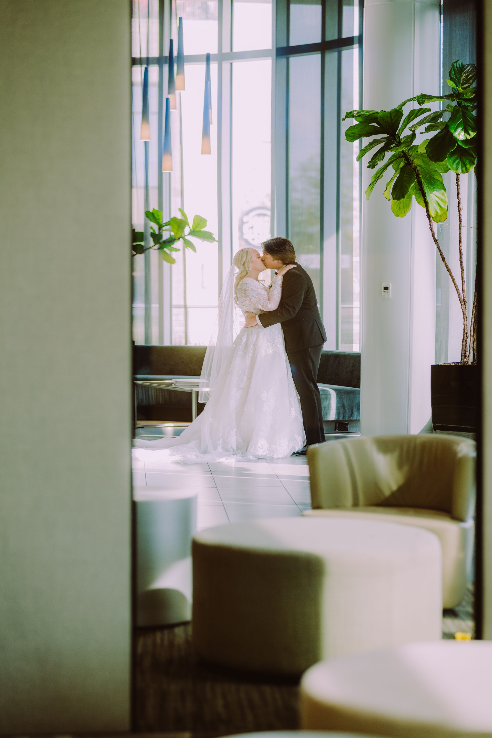 Couple kissing in front of a window, reflected in a mirror. Bride wears a white dress; groom wears a suit. Modern interior.
