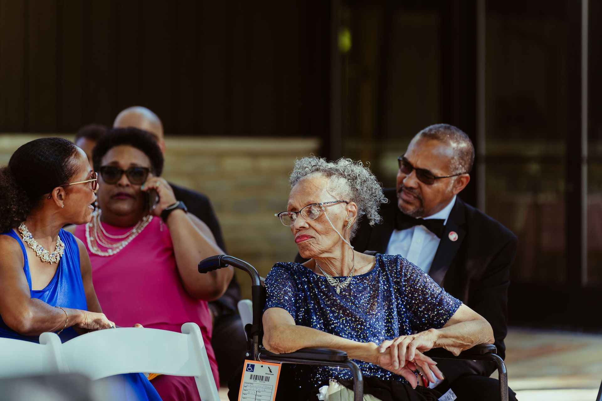 An elderly woman in a wheelchair is sitting in a chair with other people at a wedding.