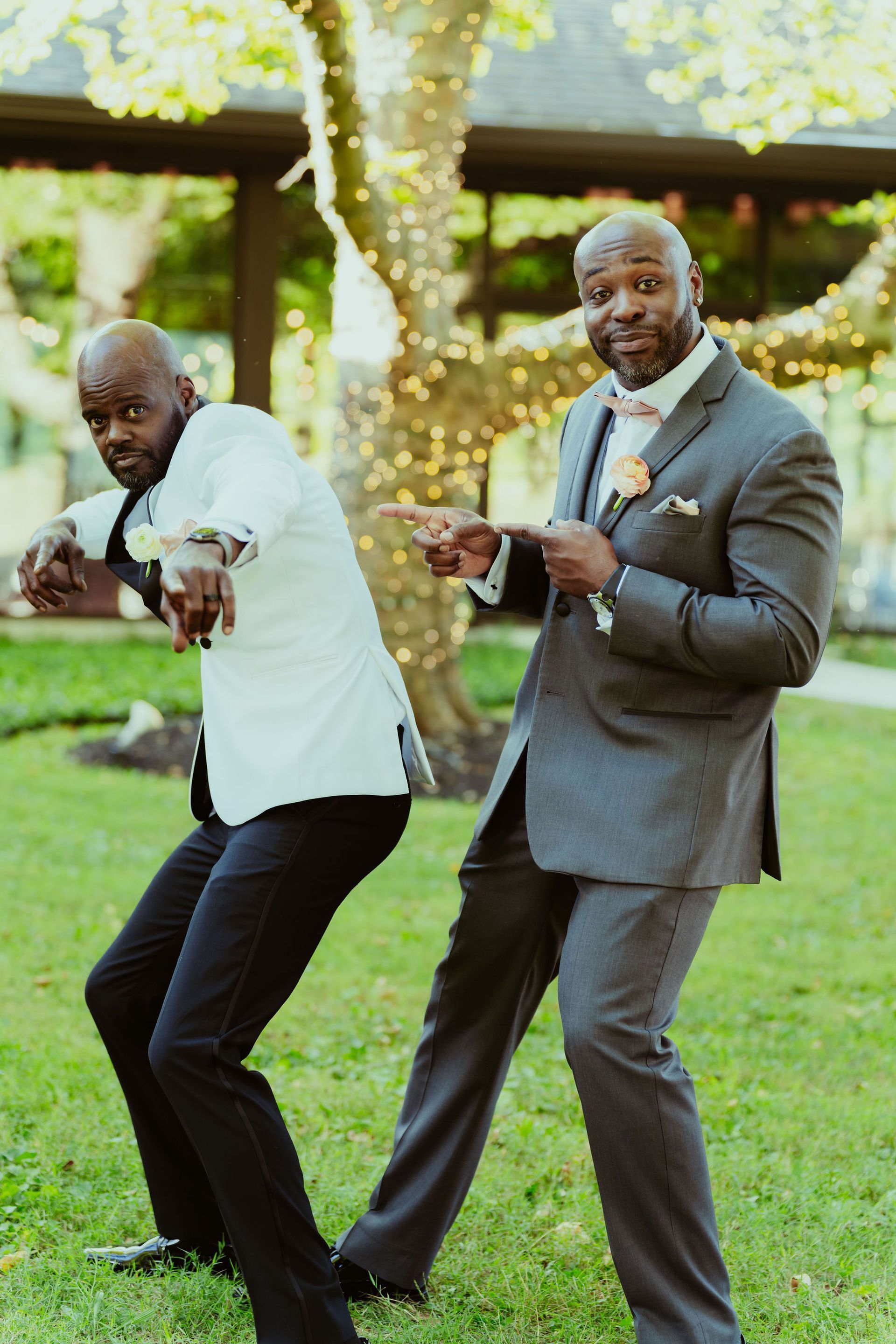 Two men in suits are standing next to each other in a park and pointing at the camera.
