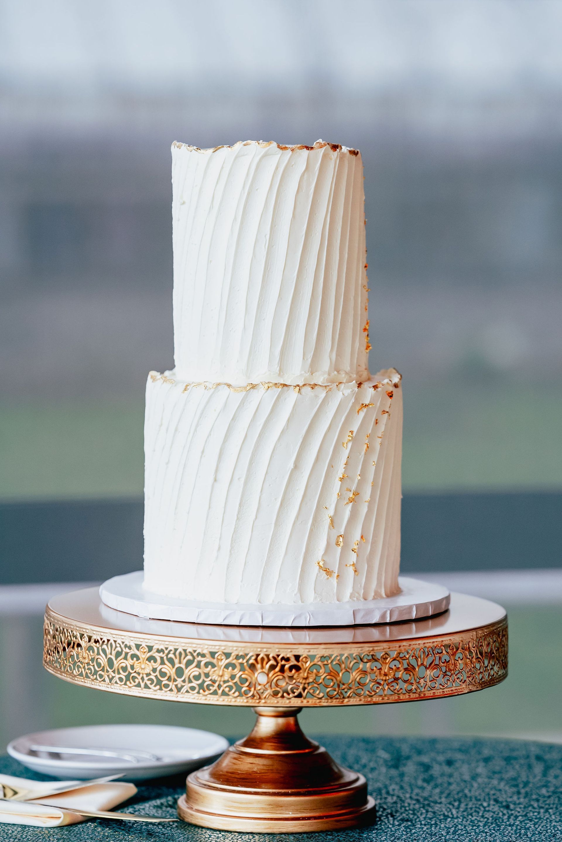 A white wedding cake is sitting on top of a gold cake stand.