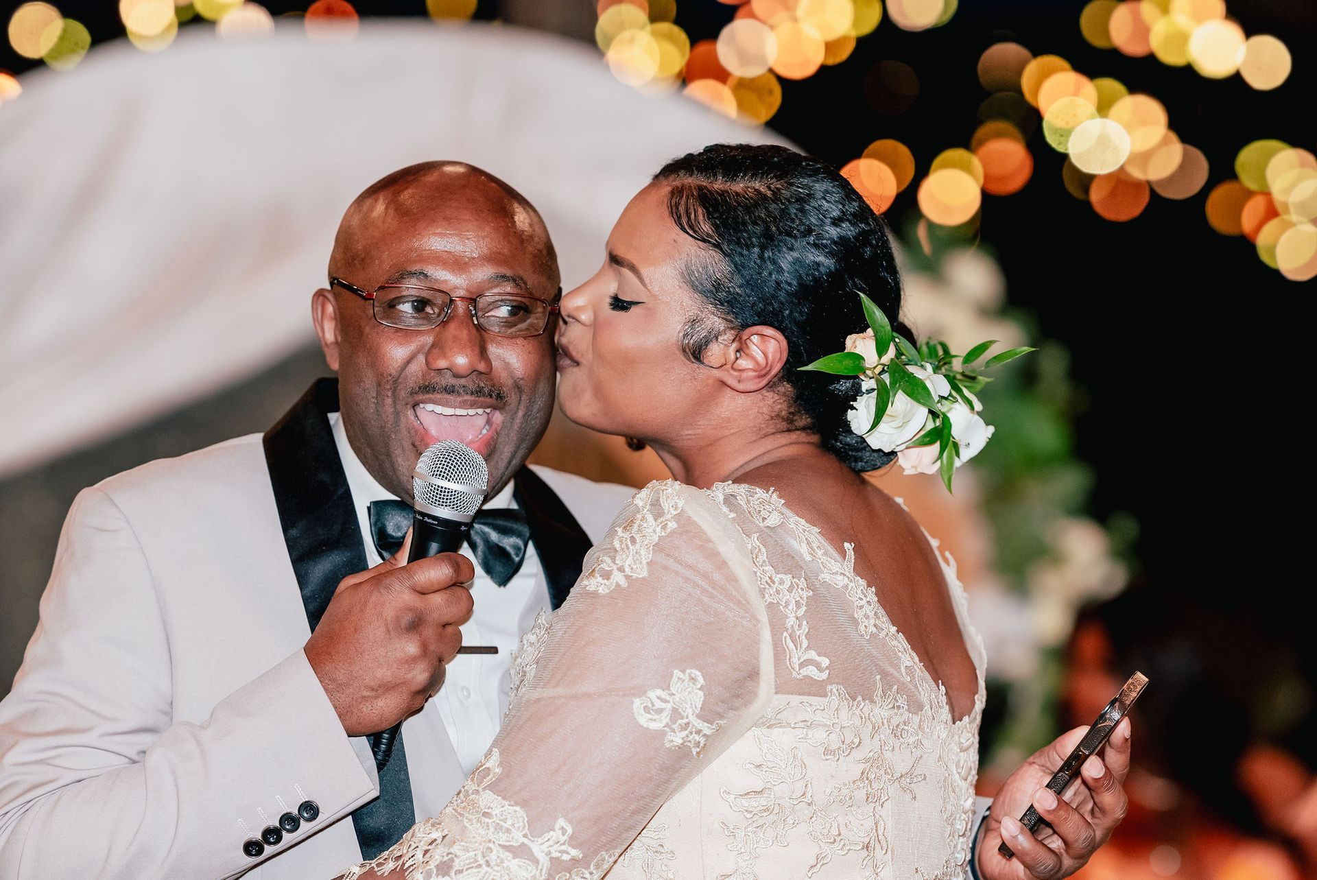 A bride is kissing her groom on the cheek while holding a microphone.