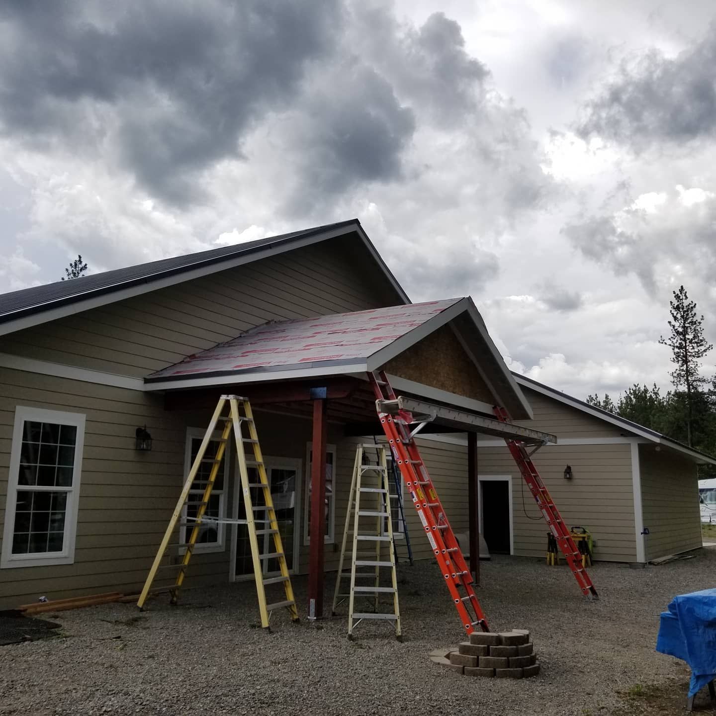 Building under construction with ladders, tan siding, and a partially built red roof, under a cloudy sky.