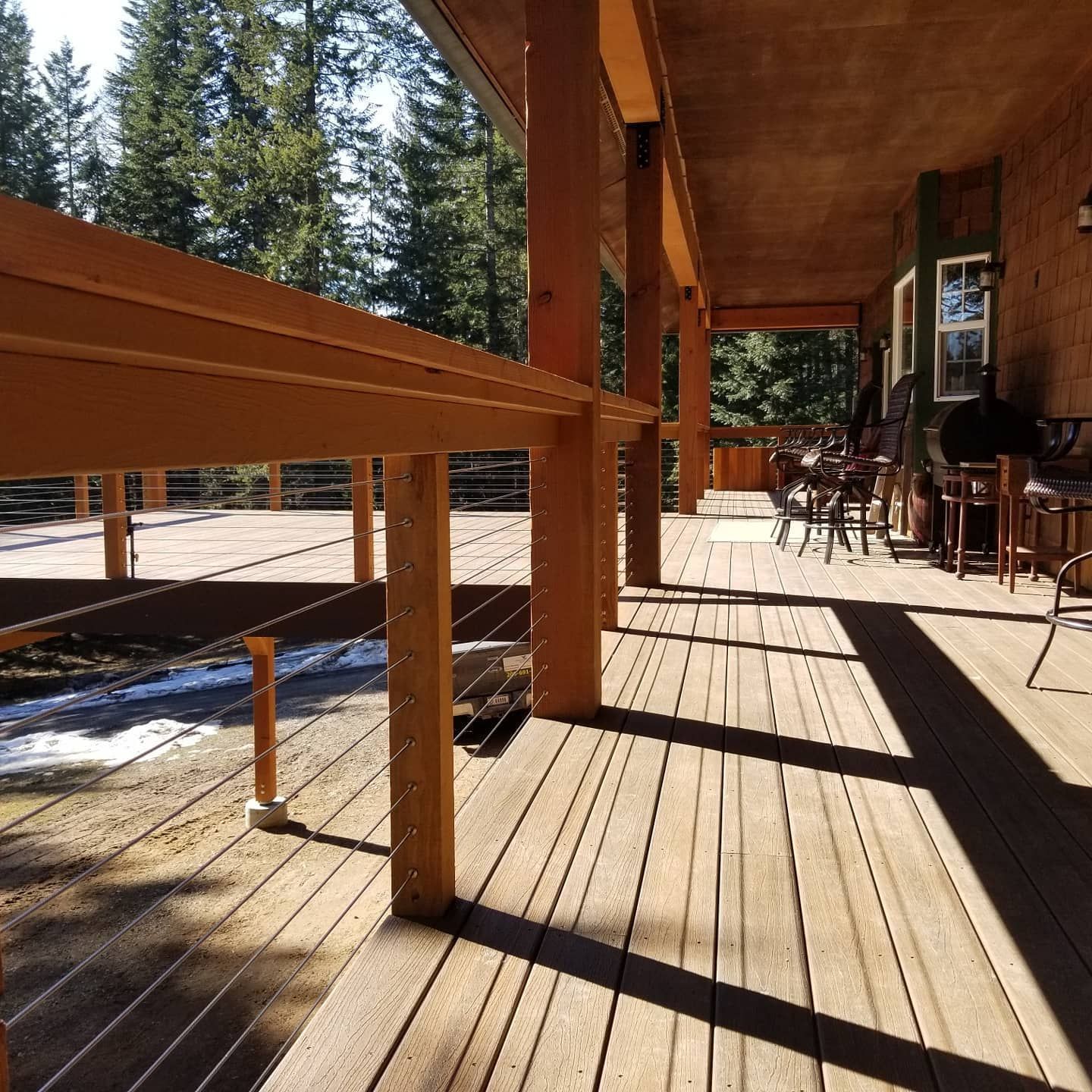 Wooden deck with cable railings and wooden posts. Shadows cast by the sun. Seating area in the background.