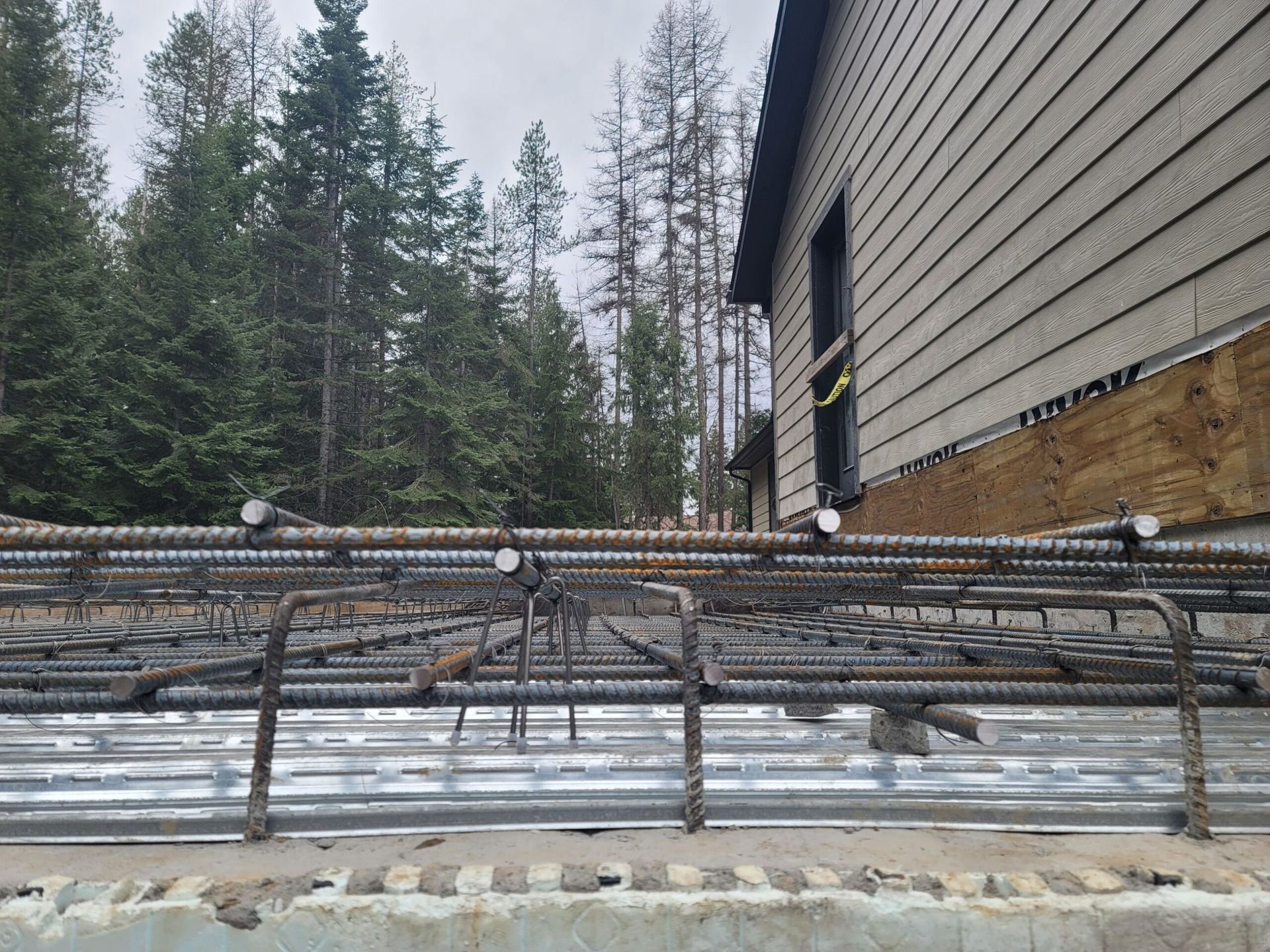 Rebar framework prepared on corrugated metal for a concrete foundation, next to a wood-sided building.