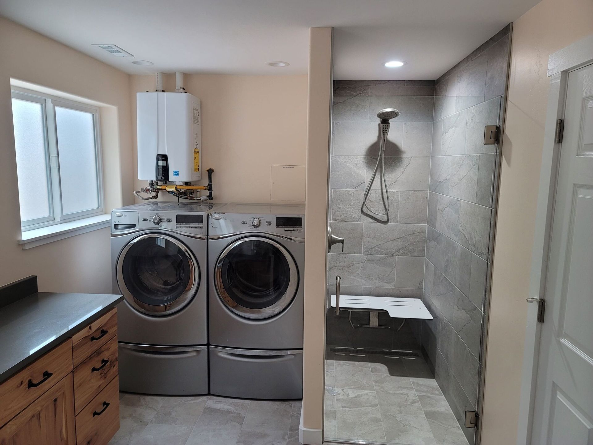 Laundry room with washer/dryer, shower, cabinetry. Gray appliances, tiled shower, window, door, and a wall-mounted water heater.