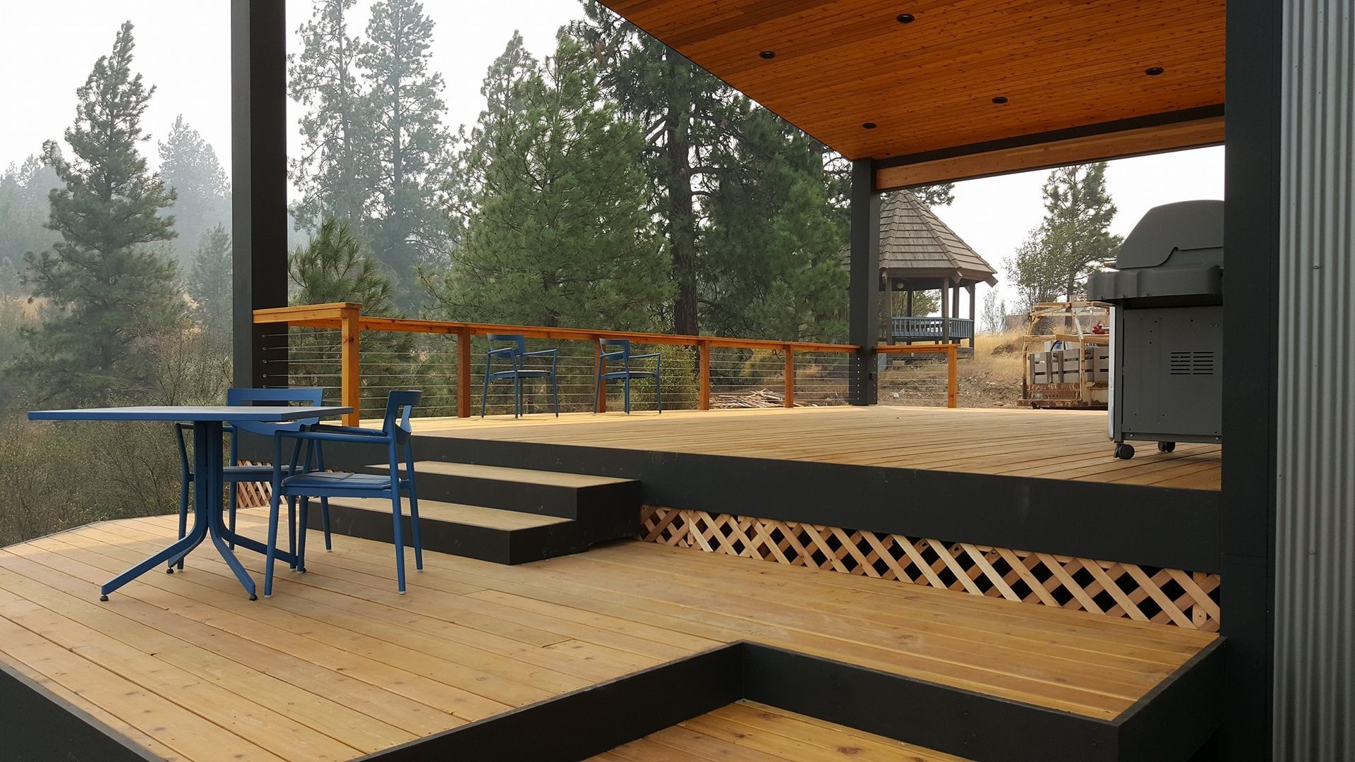 Wooden deck with blue table/chairs, grilling area, overlooking forest.