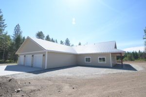 Tan, modern building with three-car garage and attached single-story section with porch. Gravel driveway, bright sky.