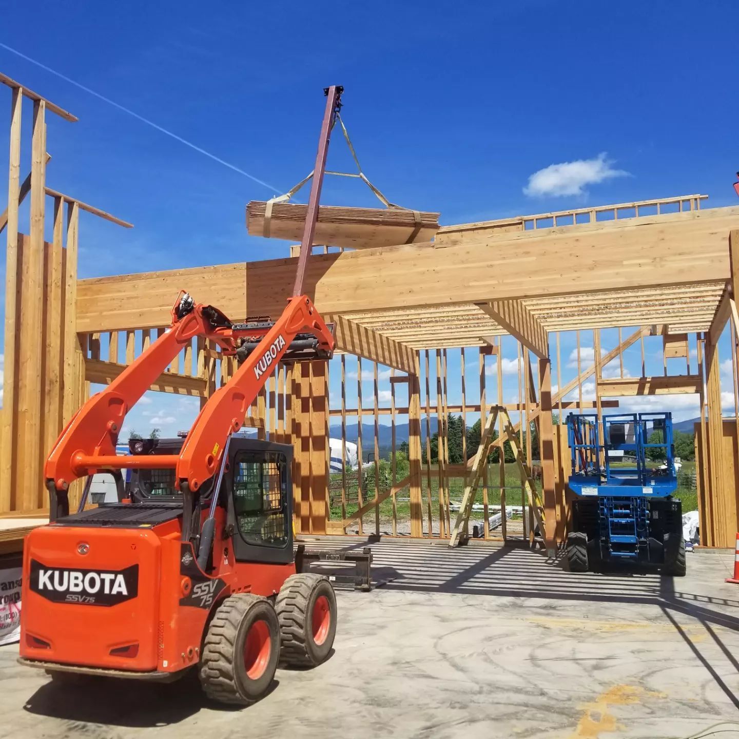 Orange Kubota skid steer lifting lumber at a construction site. Blue lift in the background. Sunny day.